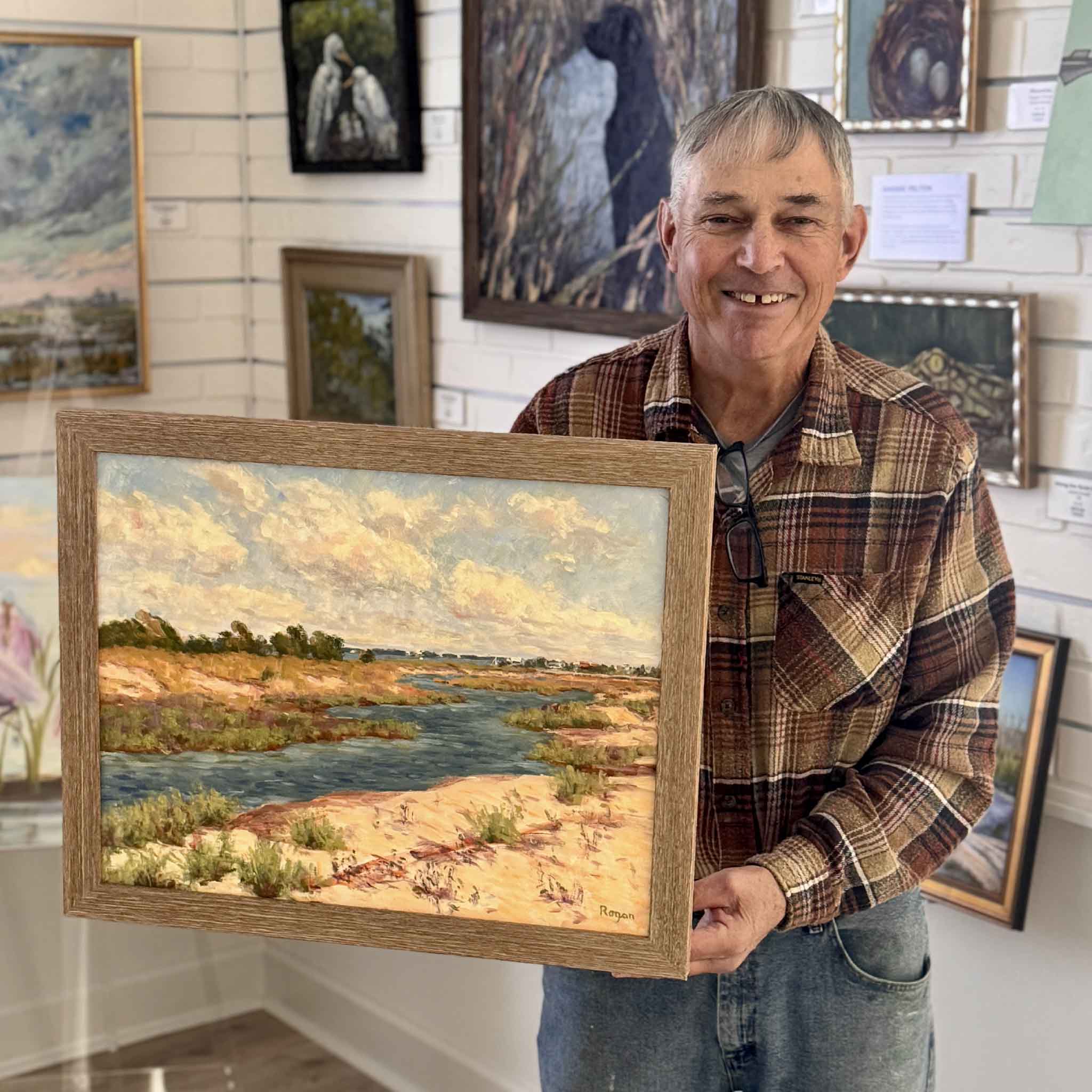 Local artist Michael Rogan holding his original oil painting Blue Creek, inspired by Huntington Beach State Park, professionally framed at Gray Man Gallery in Pawleys Island, South Carolina.