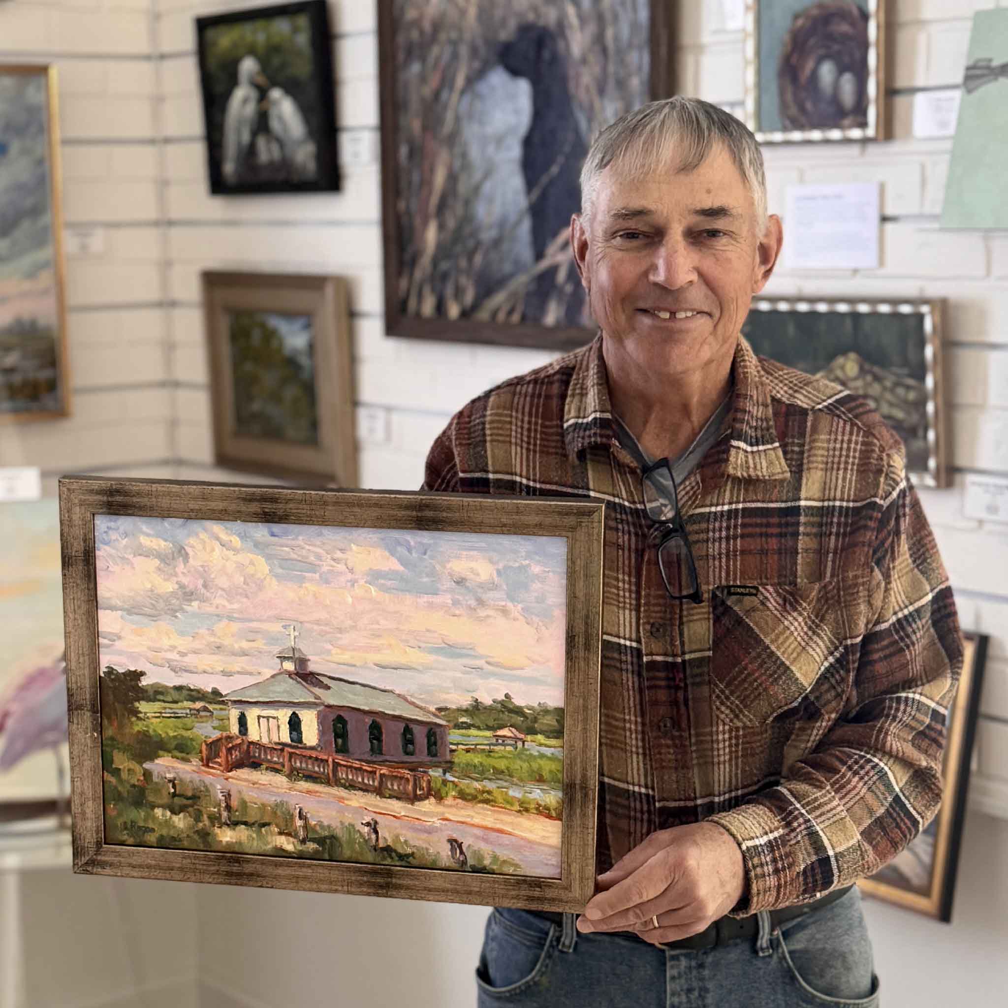 Local artist Michael Rogan at Gray Man Gallery holding his original oil painting titled Chapel on the Creek, depicting the Pawleys Island chapel surrounded by marsh and creek in soft evening light.