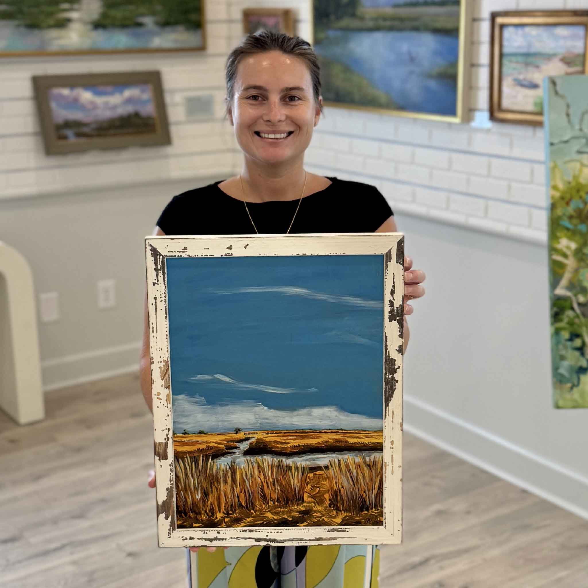 Kyra holds Autumn Sun at Clambank by Maggie Pelton inside Gray Man Gallery in Pawleys Island, South Carolina. This framed original acrylic painting depicts the golden marshes of North Inlet beneath a brilliant blue autumn sky and is presented in a distressed white wood frame that complements the warm Lowcountry landscape.