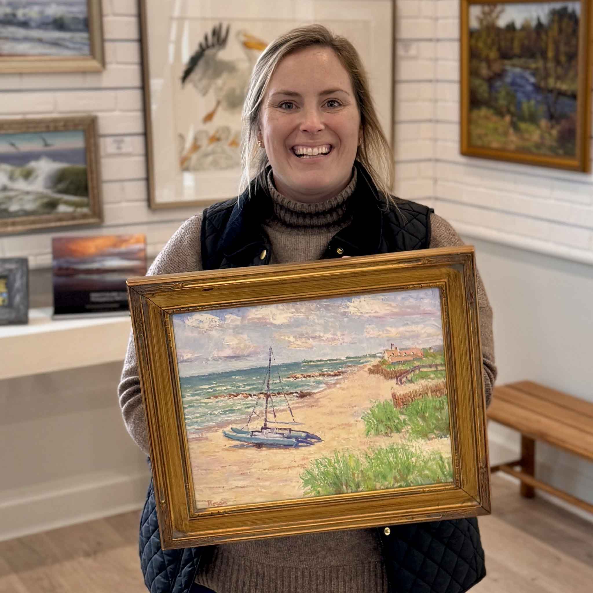 Gray Man Gallery staff member Bordy holding “Catamaran on Pawleys” original oil painting by local artist Michael Rogan, featuring a catamaran on Pawleys Island beach with dunes, sea grass, and coastal sky, displayed inside Gray Man Gallery in Pawleys Island, South Carolina