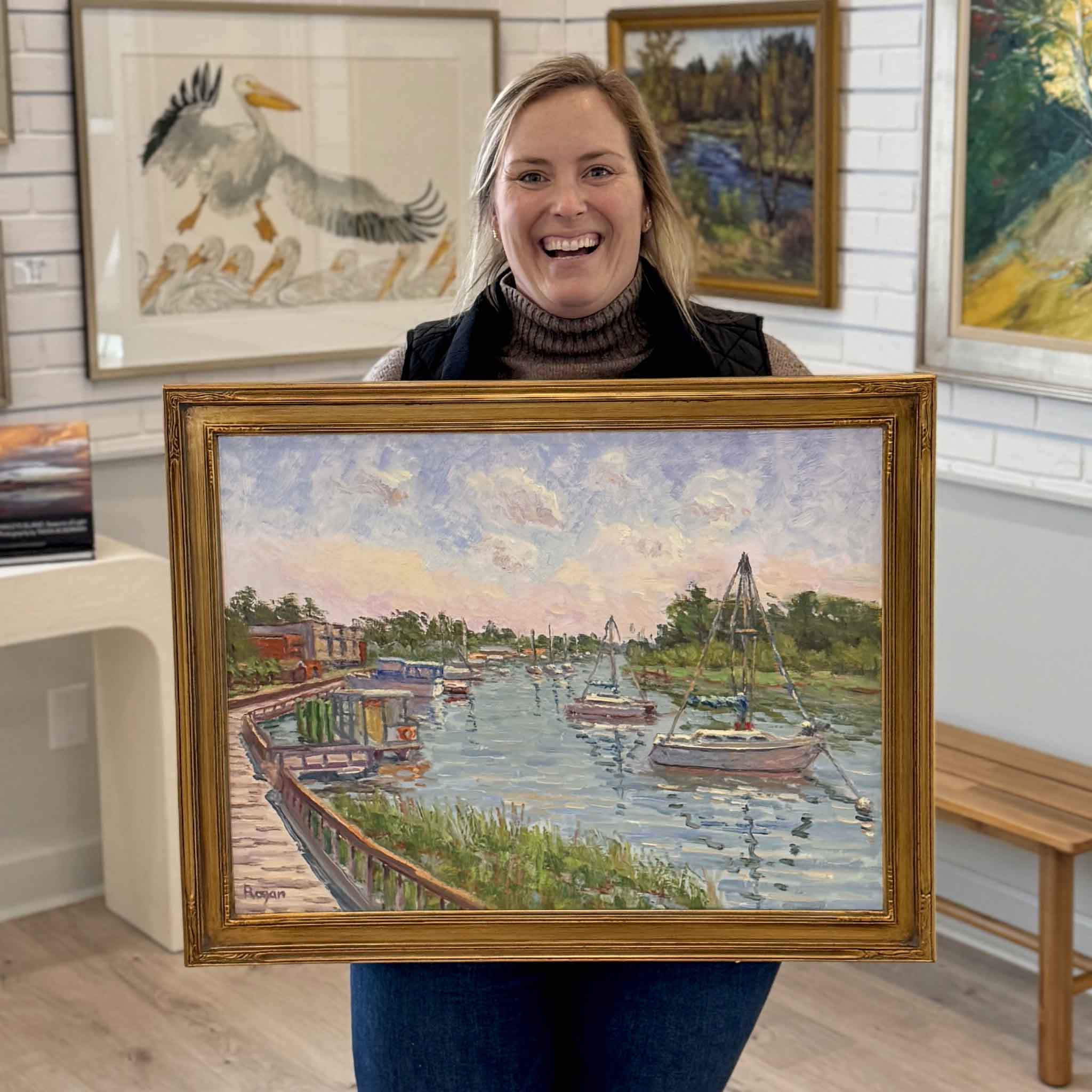 Bordy holding “Georgetown Harbor Walk,” an original oil painting by local artist Michael Rogan at Gray Man Gallery in Pawleys Island, South Carolina, featuring boats along the Georgetown waterfront and Lowcountry harbor scenery