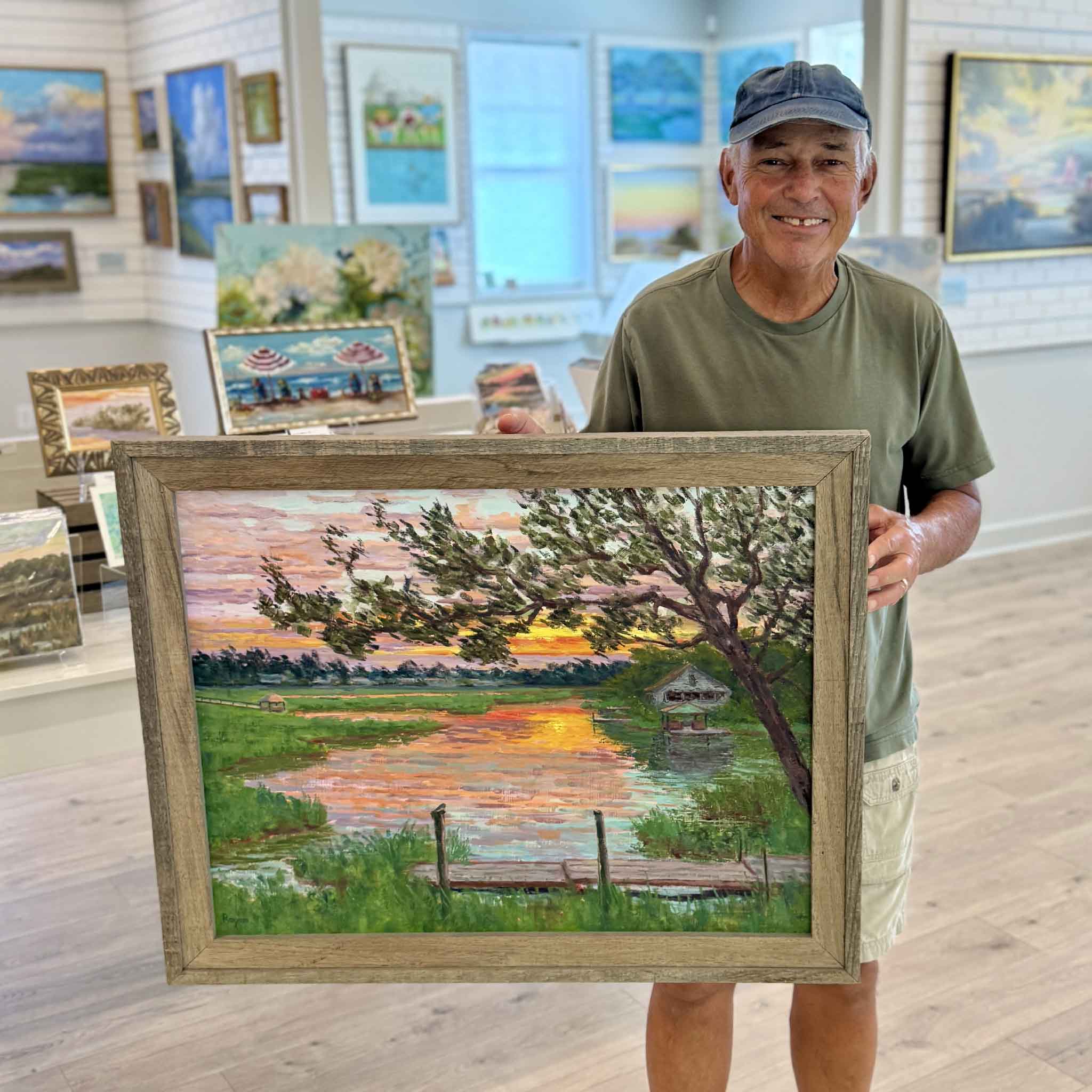 Lowcountry artist Michael Rogan holds his original en plein air oil painting Fading Light inside Gray Man Gallery in Pawleys Island, South Carolina. Painted on location in South Litchfield, the framed landscape depicts a glowing marsh at sunset with tidal creeks, a historic rice trunk, and reflections of the evening sky.