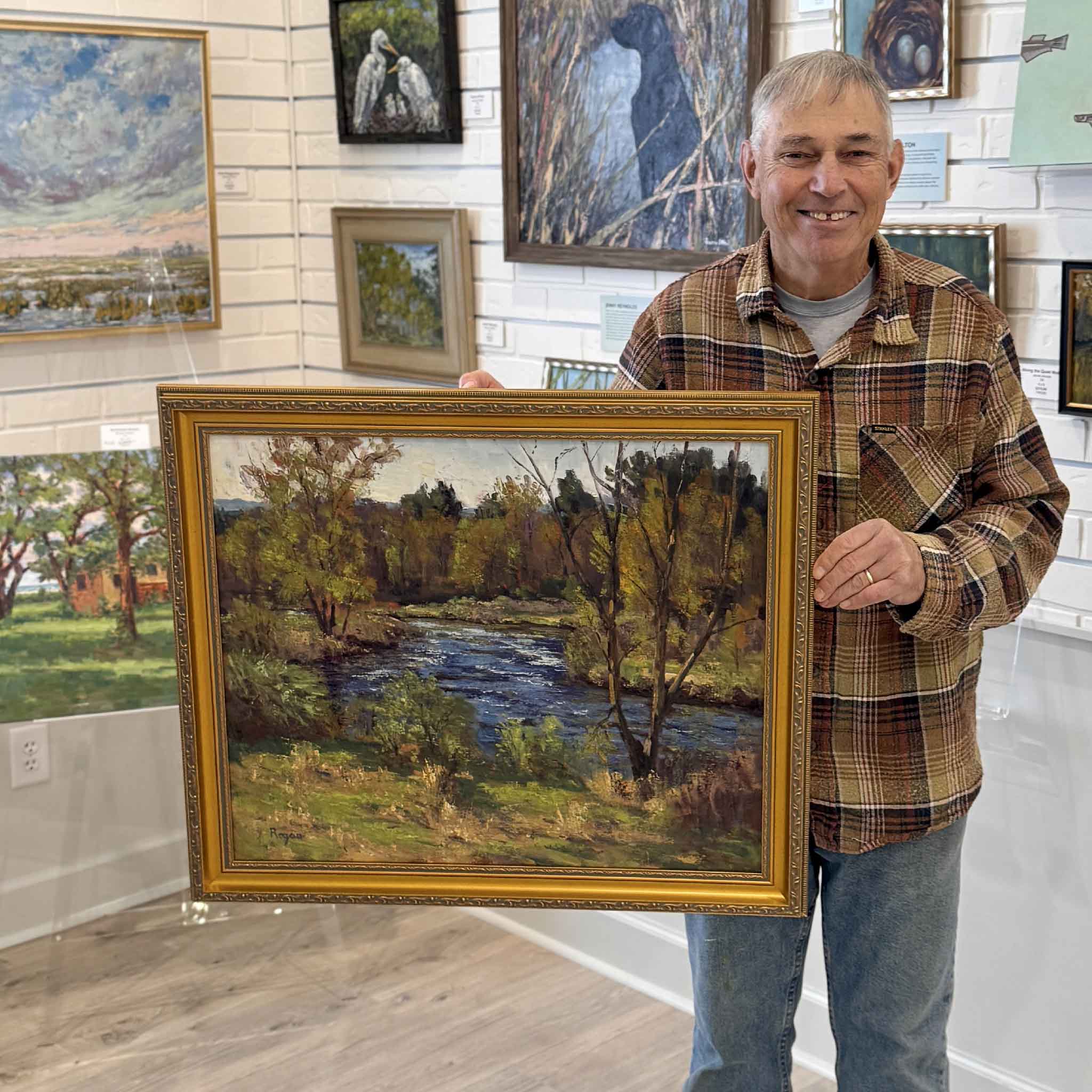 Local artist Michael Rogan holding his original oil painting Quiet Bend of the River inside Gray Man Gallery, showing the scale and framing of the 24x20 landscape artwork.