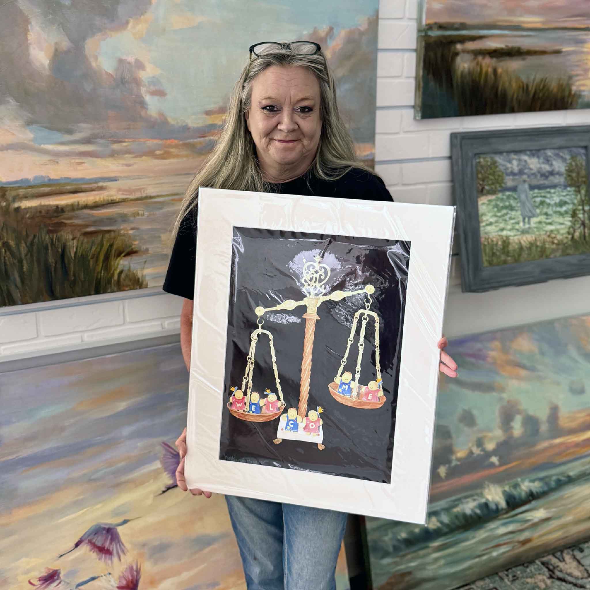 Artist Mimi Beaver holding her original watercolor Welcome inside Gray Man Gallery, showing the full scale of the framed artwork, with her signature whimsical figures on a balance scale, surrounded by coastal paintings in the Pawleys Island gallery setting.