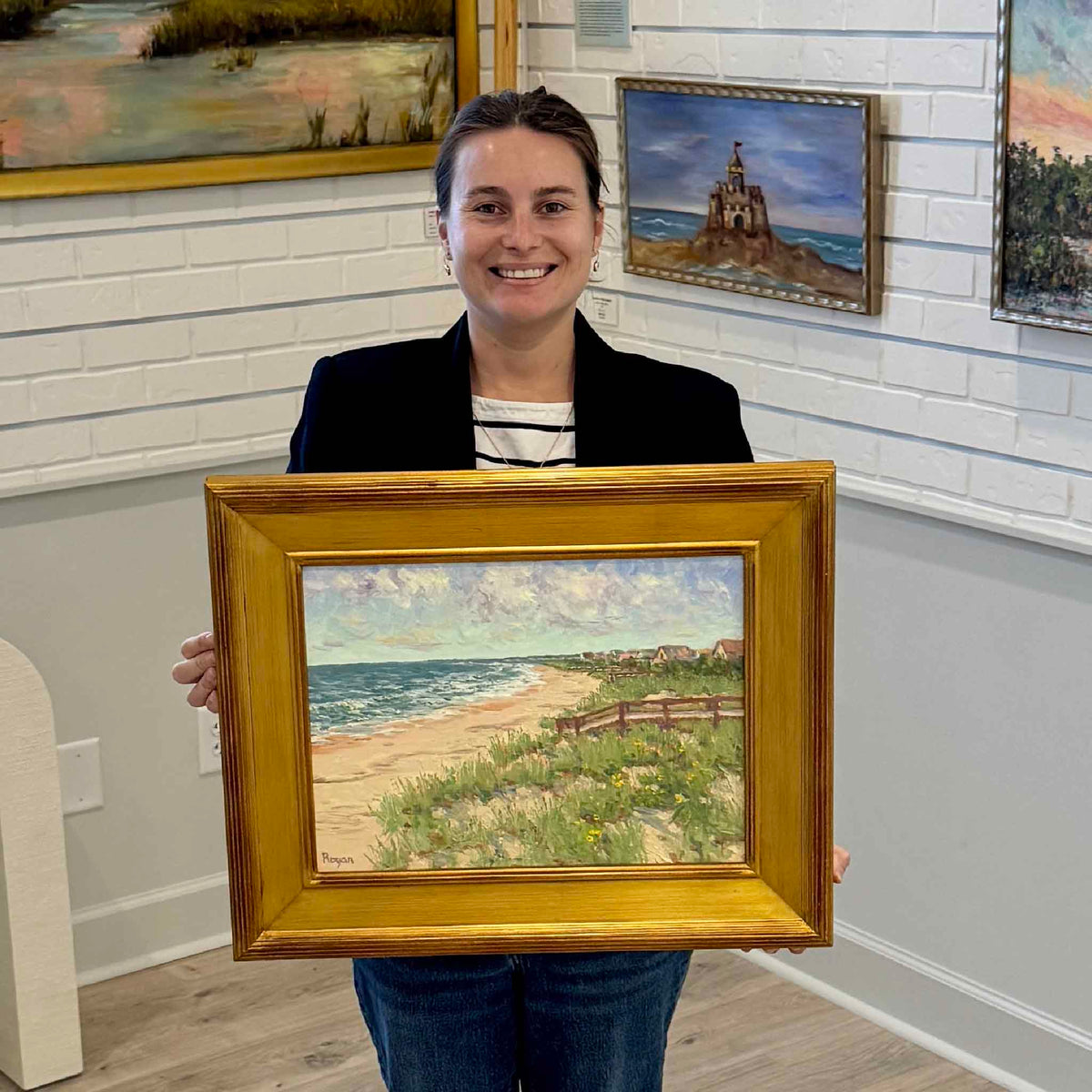Gray Man Gallery team member Kyra holding View to Inlet Point by local artist Michael Rogan inside the gallery in Pawleys Island, South Carolina. The framed coastal landscape features a sweeping Atlantic Ocean shoreline, sandy dunes, sea oats, wooden fencing, and beach cottages near the southern end of Pawleys Island. The substantial gold frame and gallery photograph provide a sense of the artwork's scale and traditional coastal presentation.