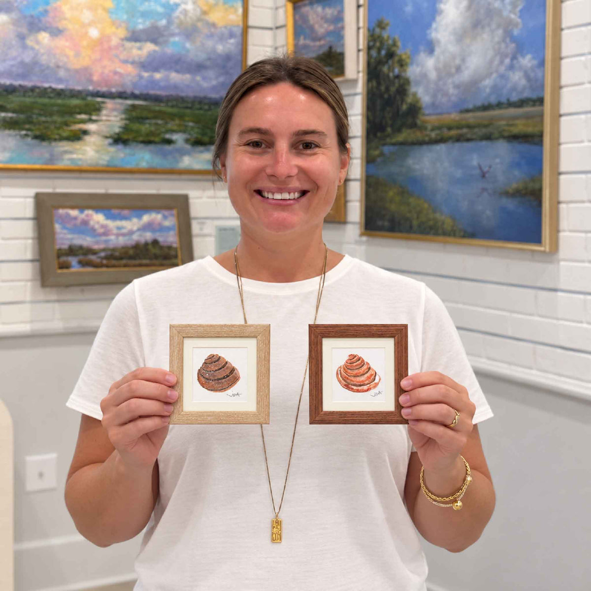Kyra holds two original Pawleys Island shell watercolor paintings by Vida Miller in light and dark wood shadowbox frames inside Gray Man Gallery in Pawleys Island, South Carolina.