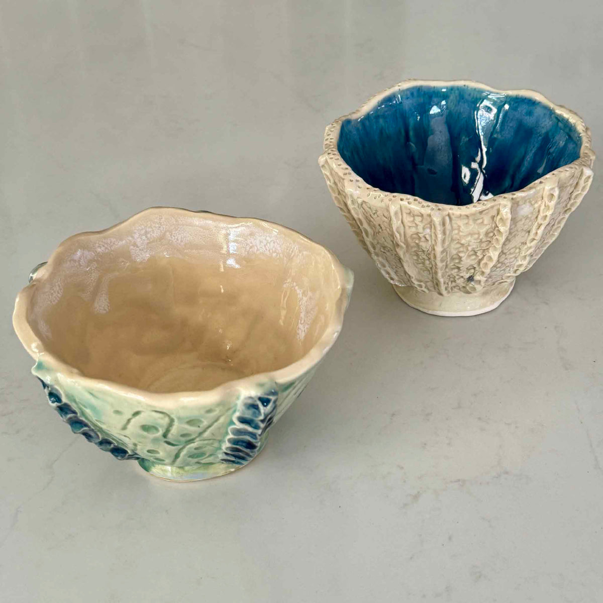 Two handmade Tidepool Texture Bowls by Pawleys Island artist and Gray Man Gallery founder Vida Miller, showing one bowl with a seafoam green textured exterior and warm cream interior and another with a sculptural ivory exterior and deep cobalt blue glaze, each inspired by the textures and colors of the South Carolina Lowcountry coast.
