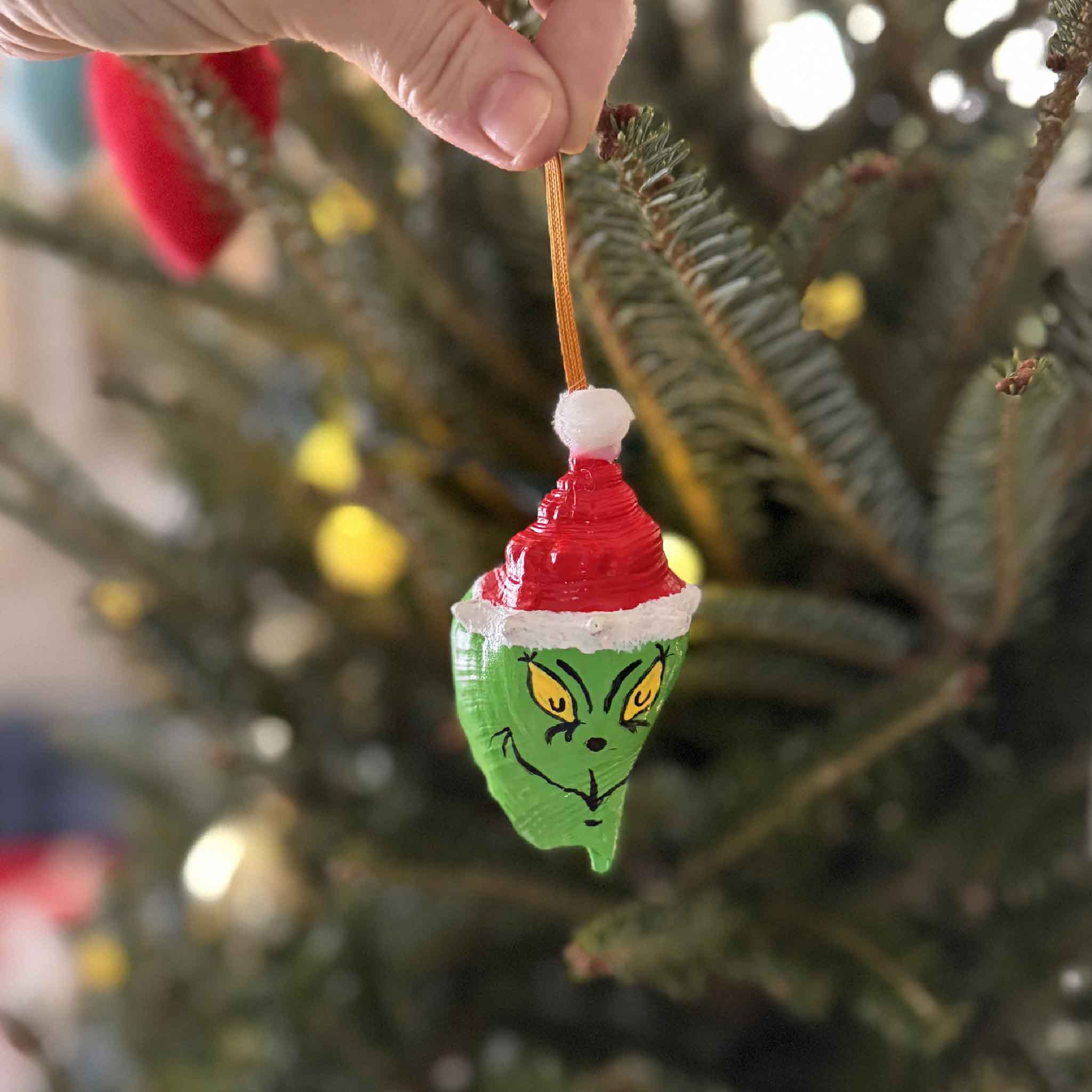 This photo shows a hand-painted Grinch conch shell ornament being hung on a Christmas tree, highlighting its bright colors and coastal charm.