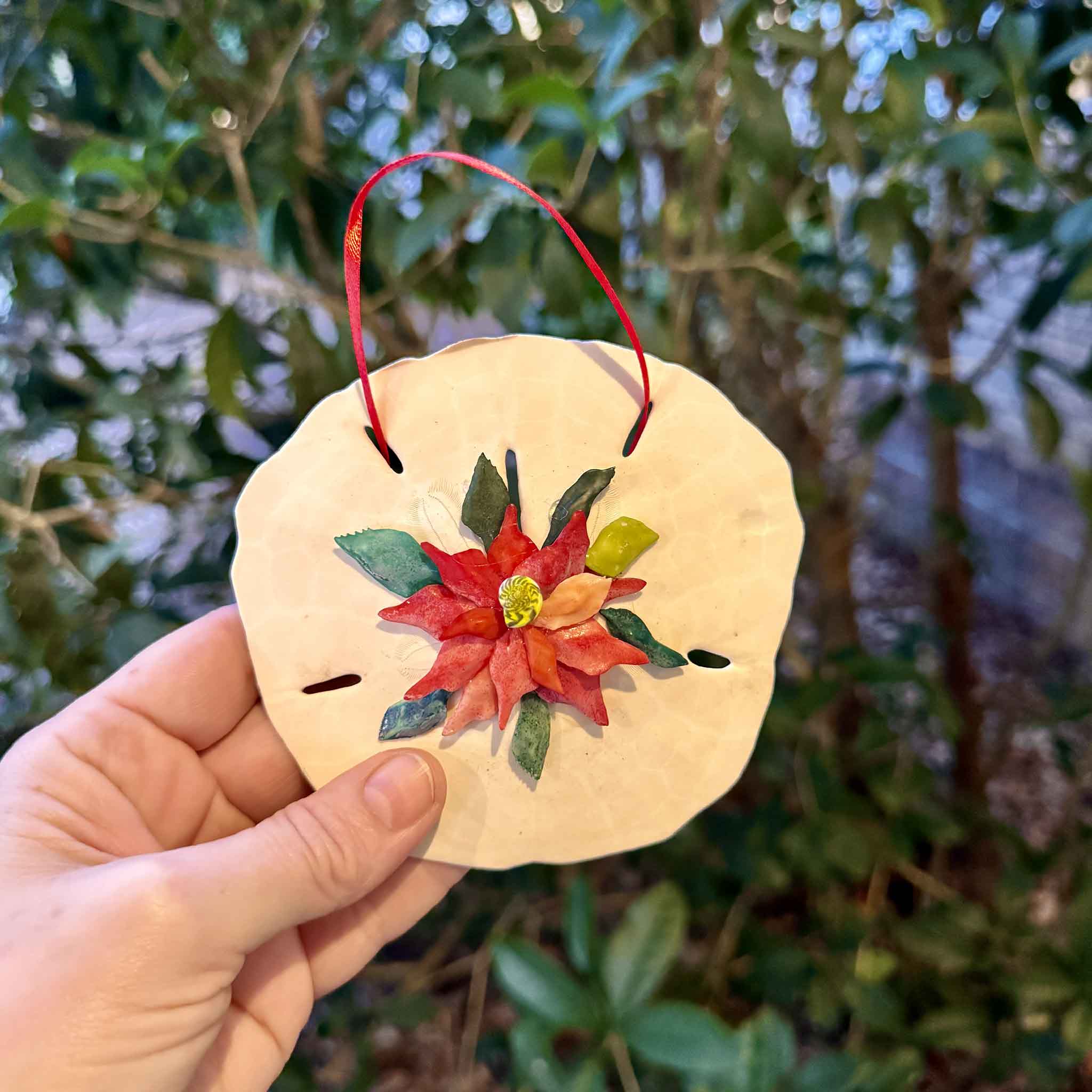 Hand holding a handmade poinsettia sand dollar ornament by Pawleys Island artist Tootsie Watkins