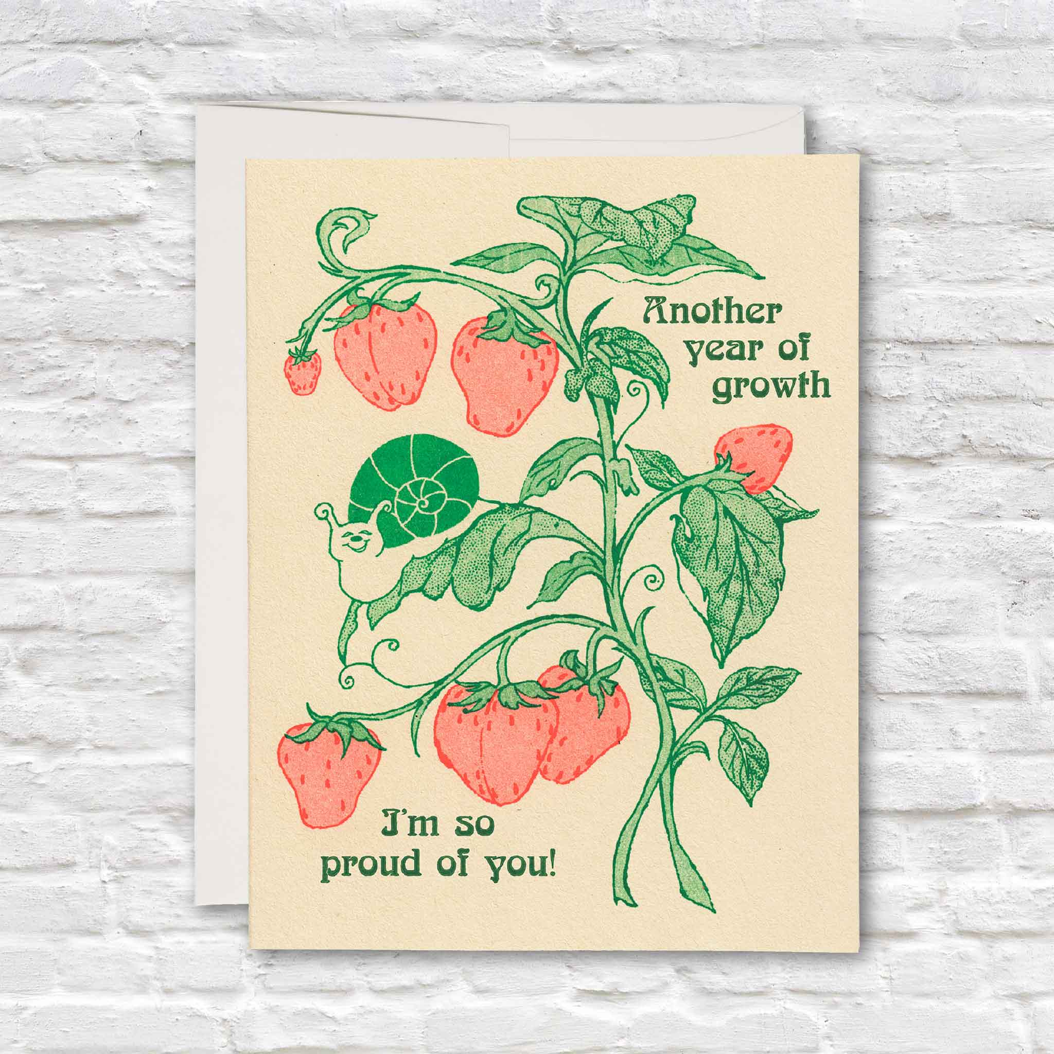Another Year of Growth greeting card illustrated by Hannah-Michelle Bayley, featuring red strawberries, green vines and a tiny snail with the message “I’m so proud of you,” available inside Gray Man Gallery in Pawleys Island, South Carolina.