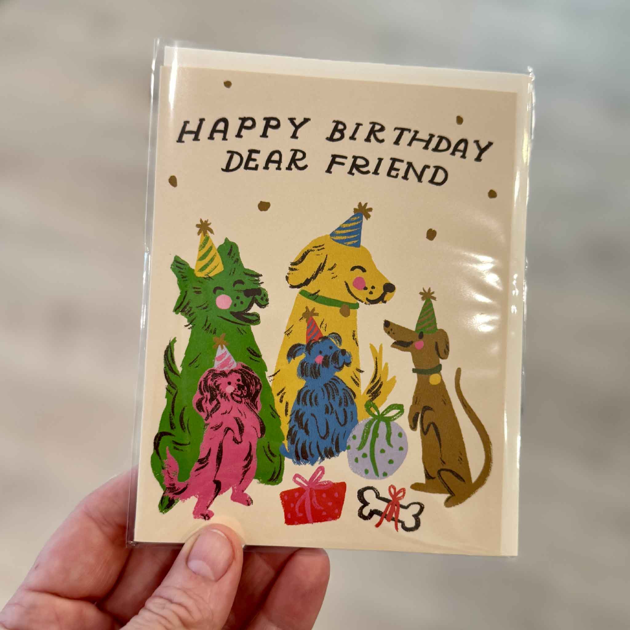 A hand holds the Happy Birthday Dear Friend greeting card by Dylan Mierzwinski inside Gray Man Gallery in Pawleys Island, South Carolina. Featuring a playful group of colorful dogs wearing birthday hats alongside presents and confetti, this blank-inside Red Cap Cards greeting card is perfect for dog lovers, best friends, family members, coworkers, and anyone celebrating a birthday with a smile.
