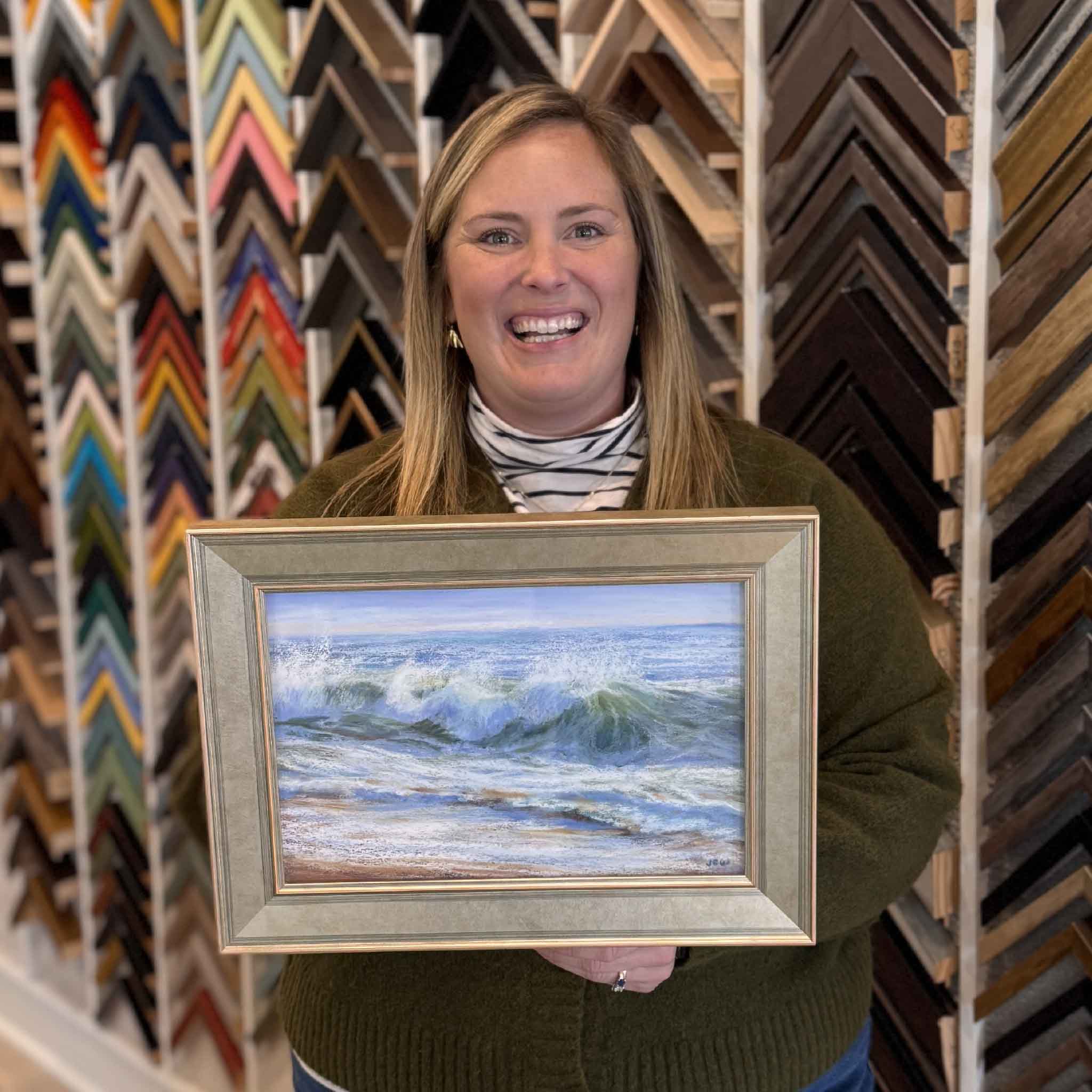 Original pastel seascape titled “Happy Dance” by local artist Joanne Willoughby, depicting rolling green-blue waves breaking along the shoreline with foamy white surf and sandy undertones, shown professionally framed and held inside Gray Man Gallery in Pawleys Island, South Carolina, photographed in the gallery to show scale, framing detail, texture, and coastal movement