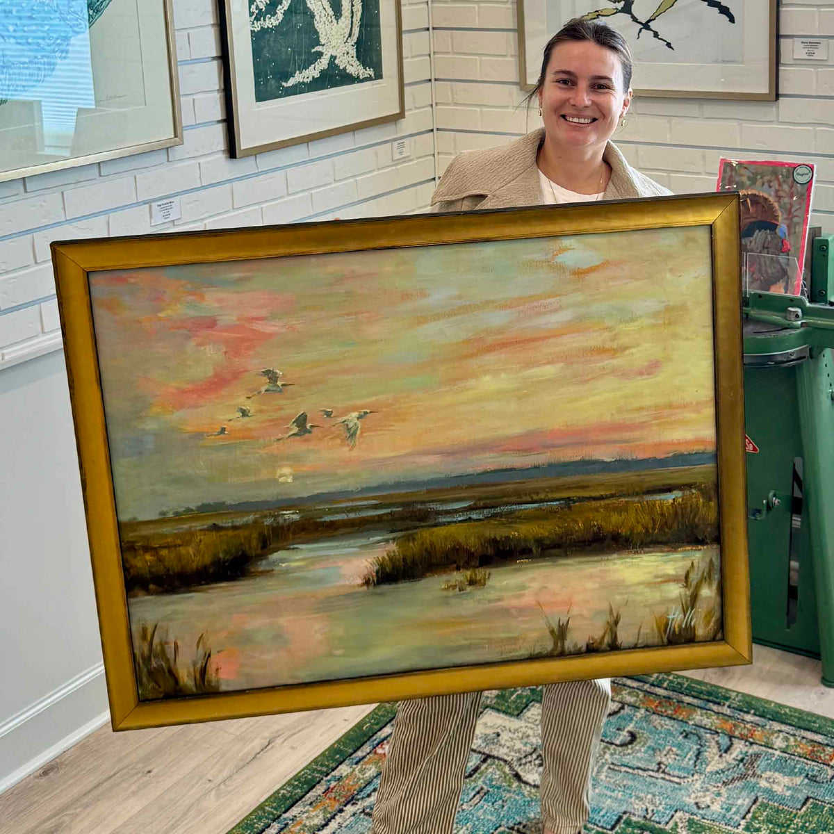 Gray Man Gallery team member holding Chasing the Last Light, a large original oil painting by local artist Helli Luck, inside Gray Man Gallery in Pawleys Island, South Carolina. The horizontal coastal landscape features birds flying across a colorful pink and golden sunset above winding tidal creeks and South Carolina Lowcountry marsh grasses. Displayed in an elegant gold frame, the photograph highlights the artwork's impressive scale.