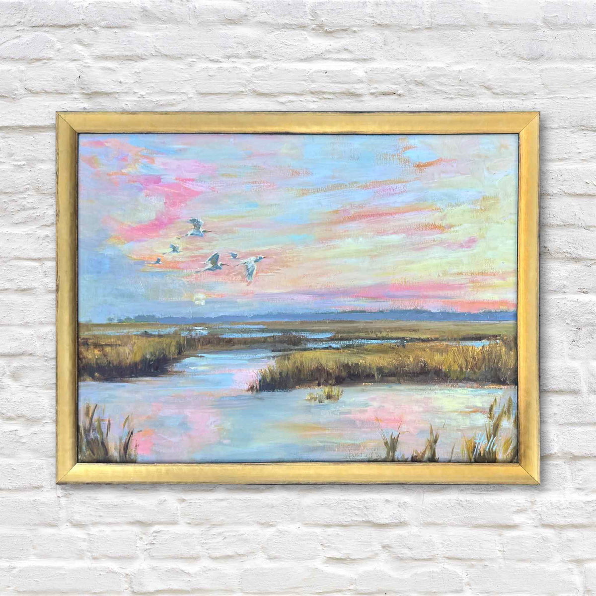 Chasing the Last Light, an original oil painting by local South Carolina artist Helli Luck, depicting coastal birds flying above a winding Lowcountry tidal creek at sunset. Soft pink, lavender, peach, and golden clouds stretch across the sky, reflecting in peaceful marsh waters surrounded by golden grasses. Presented in a classic gold frame against the white brick wall at Gray Man Gallery in Pawleys Island, South Carolina.