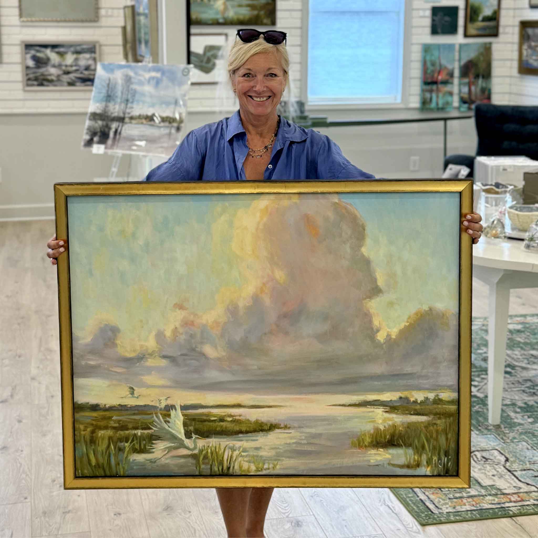 Artist Helli Luck holding Journey's End, her original oil painting depicting a luminous Lowcountry marsh scene with soaring egrets, reflective water, and dramatic pastel clouds, photographed inside Gray Man Gallery in Pawleys Island, South Carolina.