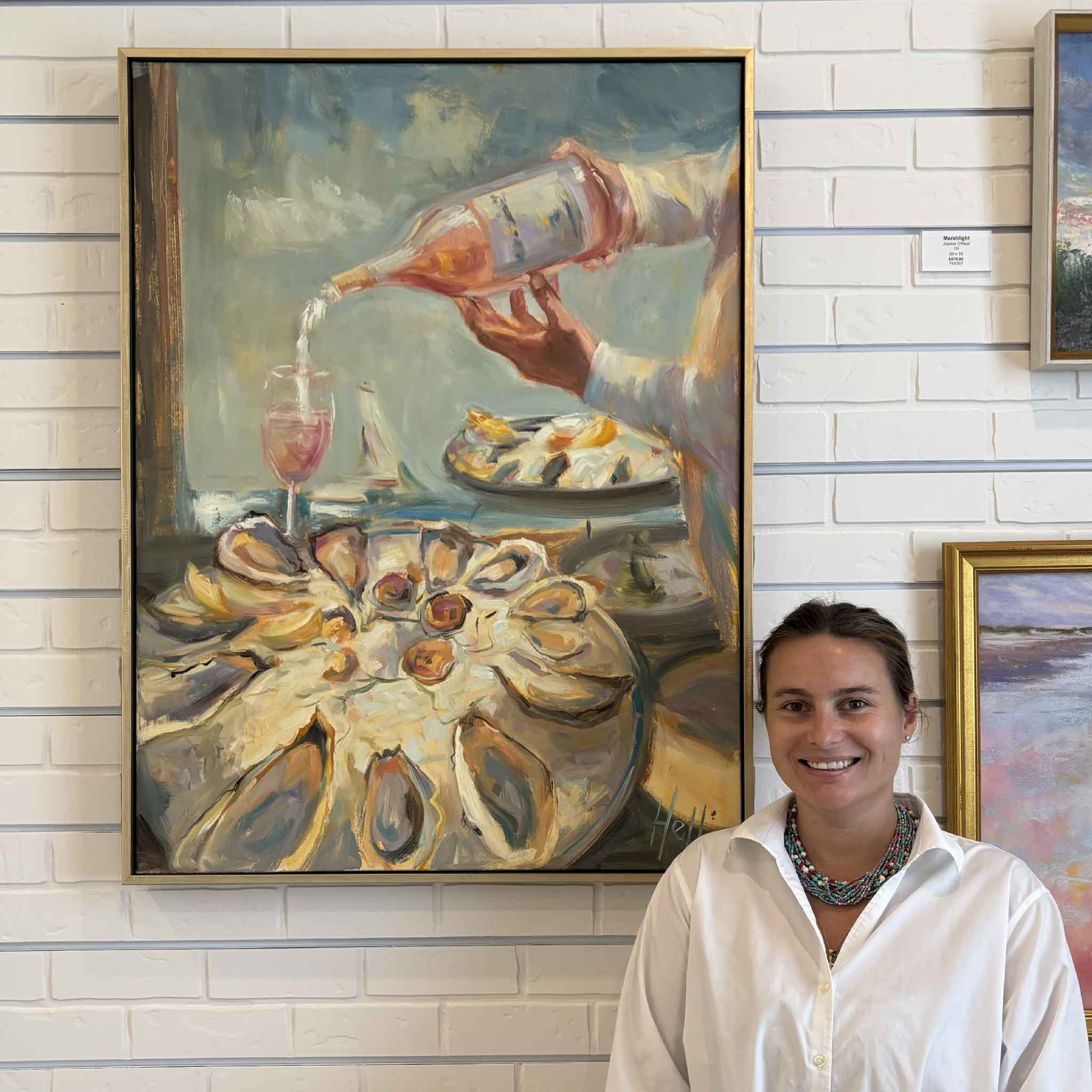 Artist Helli Luck stands beside her framed original oil painting A Taste of Summer at Gray Man Gallery in Pawleys Island, South Carolina. The large impressionistic oil painting features a platter of fresh oysters, rosé being poured into a wine glass, and soft coastal light, showcasing Helli Luck's signature expressive brushwork and contemporary Lowcountry style.