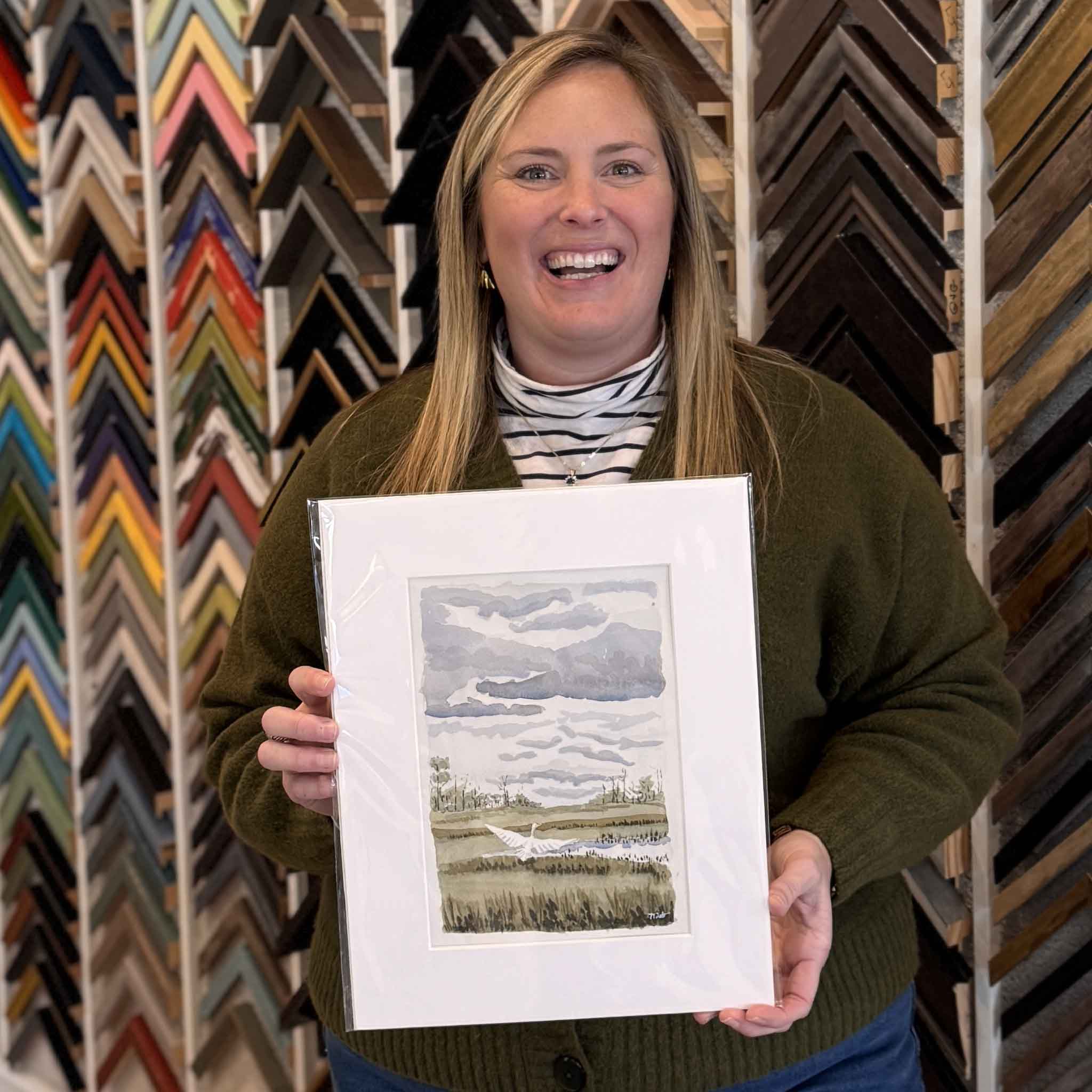 Bordy holding local artist Maggie Pelton's original watercolor Heron Taking Flight at Gray Man Gallery in Pawleys Island, South Carolina. The artwork captures a heron in mid-flight above a quiet Lowcountry marsh and measures 7x10 inches before matting. It is professionally matted to fit a standard 11x14 frame, providing an easy, ready-to-frame presentation and clear reference for scale.