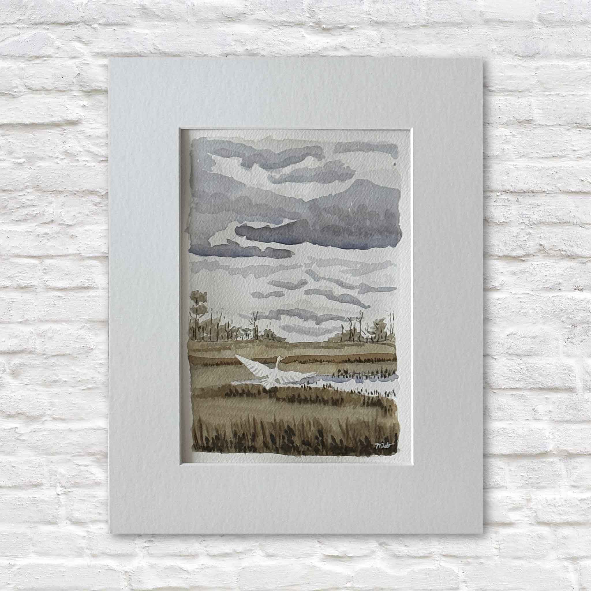 Original watercolor titled Heron Taking Flight by local artist Maggie Pelton, depicting a white heron lifting into flight over a Lowcountry marsh beneath layered gray-blue coastal skies. The 7x10 inch artwork is professionally matted to fit a standard 11x14 frame and is shown matted against a white brick backdrop at Gray Man Gallery in Pawleys Island, South Carolina.