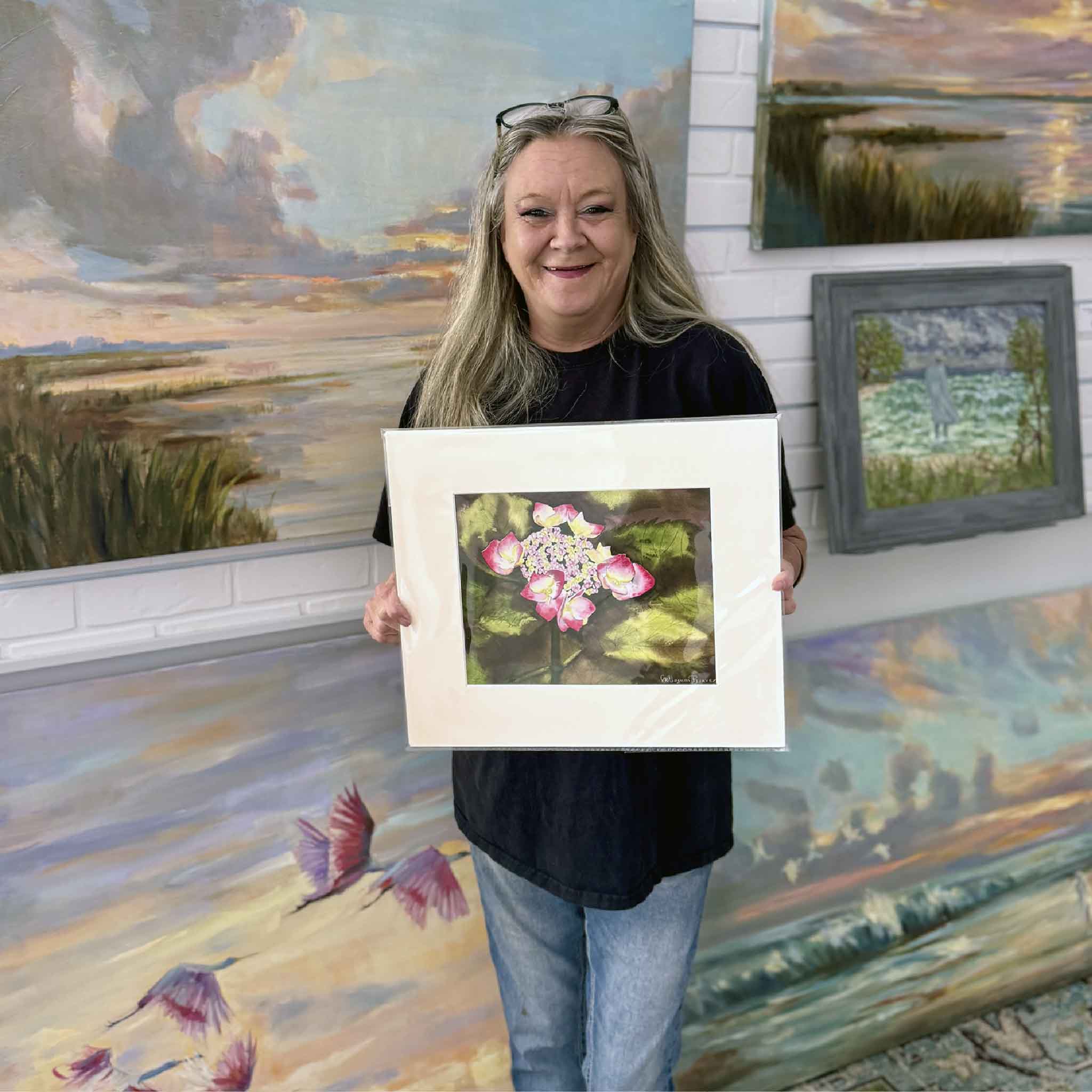 A Gray Man Gallery team member holds Hydrangea 4 by Mimi Beaver, an original watercolor featuring a pale lavender hydrangea cluster surrounded by pink-tipped petals and layered green leaves, donated to benefit Salty Dogs Senior Animal Rescue.