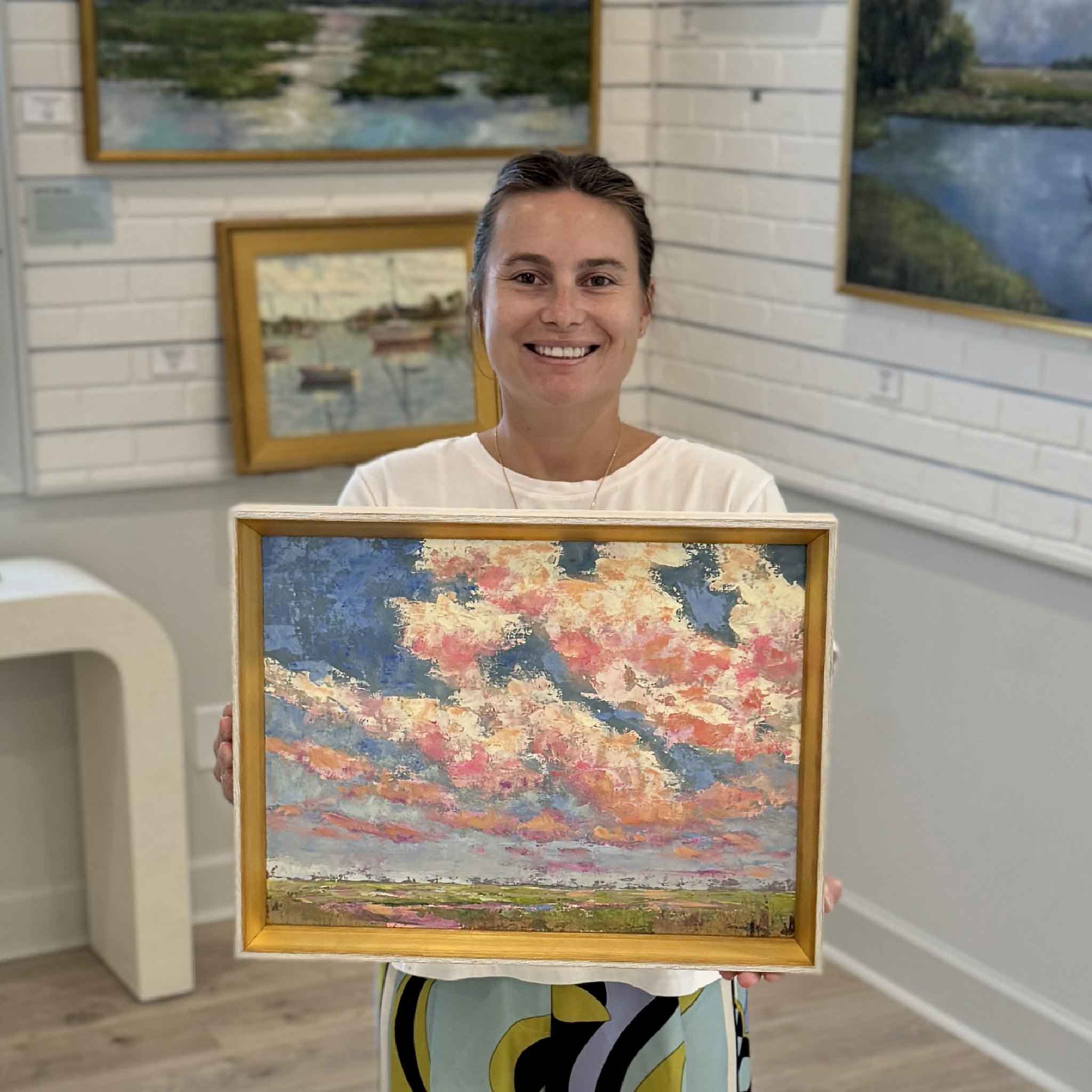 Kyra holding Beneath the Blushing Sky, an original oil painting by Pawleys Island artist Jeanne O’Neal, inside Gray Man Gallery in Pawleys Island, South Carolina, showing the scale of the framed 16 by 12 inch Lowcountry landscape. The colorful coastal painting features expressive pink, coral and cream clouds sweeping across a blue evening sky above the marsh and is finished in Jeanne O’Neal’s signature gold frame.