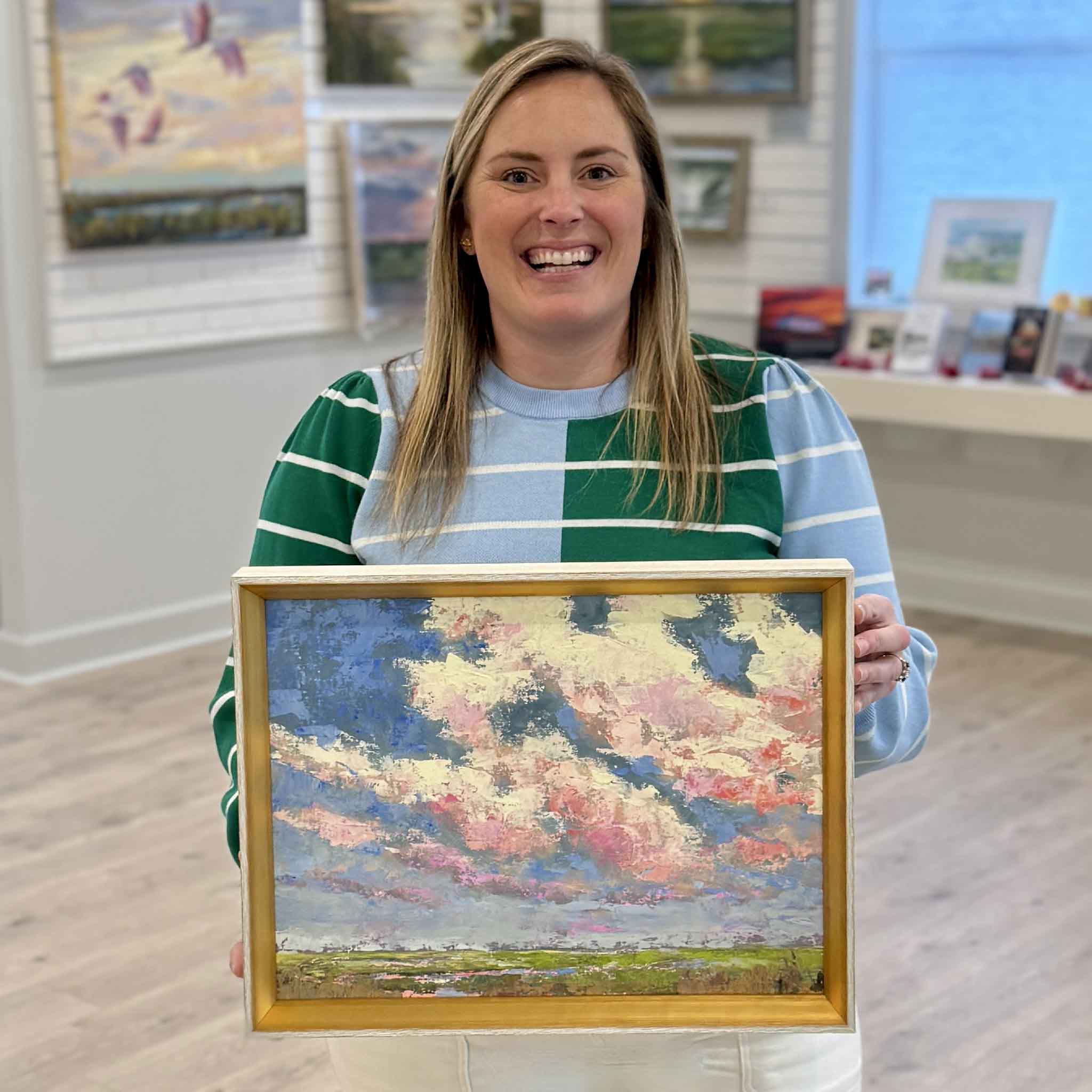 Gray Man Gallery staff member Bordy holding Beneath the Blushing Sky original oil painting by Jeanne O’Neal framed in gold showing Lowcountry sunset marsh scene in Pawleys Island South Carolina