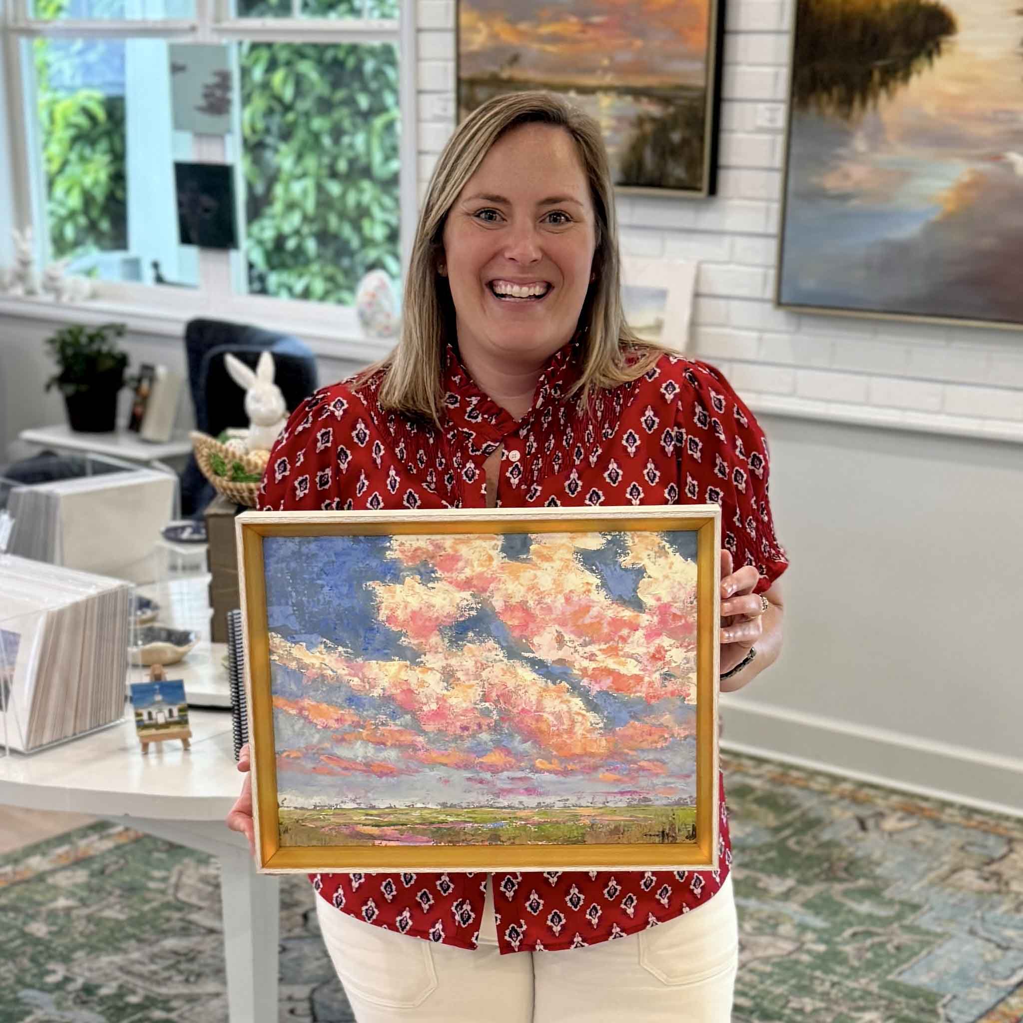 Gray Man Gallery staff member Bordy holding Beneath the Blushing Sky original oil painting by Jeanne O’Neal framed in gold showing Lowcountry sunset marsh scene in Pawleys Island South Carolina