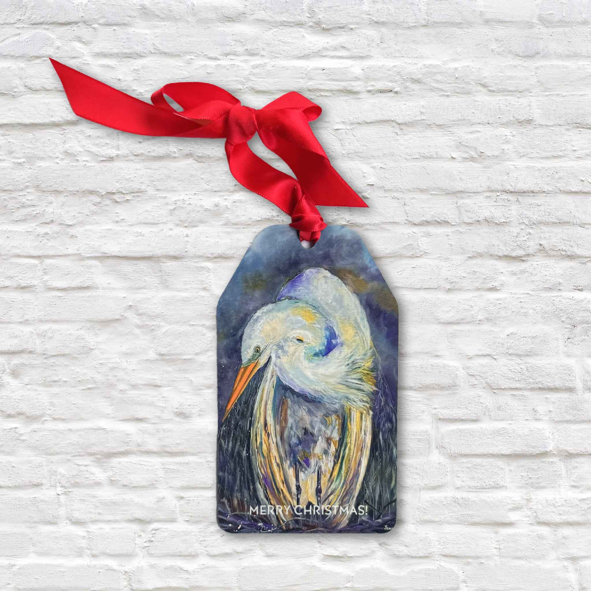 Jeanne O’Neal Coastal Christmas Ornament featuring an egret painting with red ribbon, hanging against white brick.