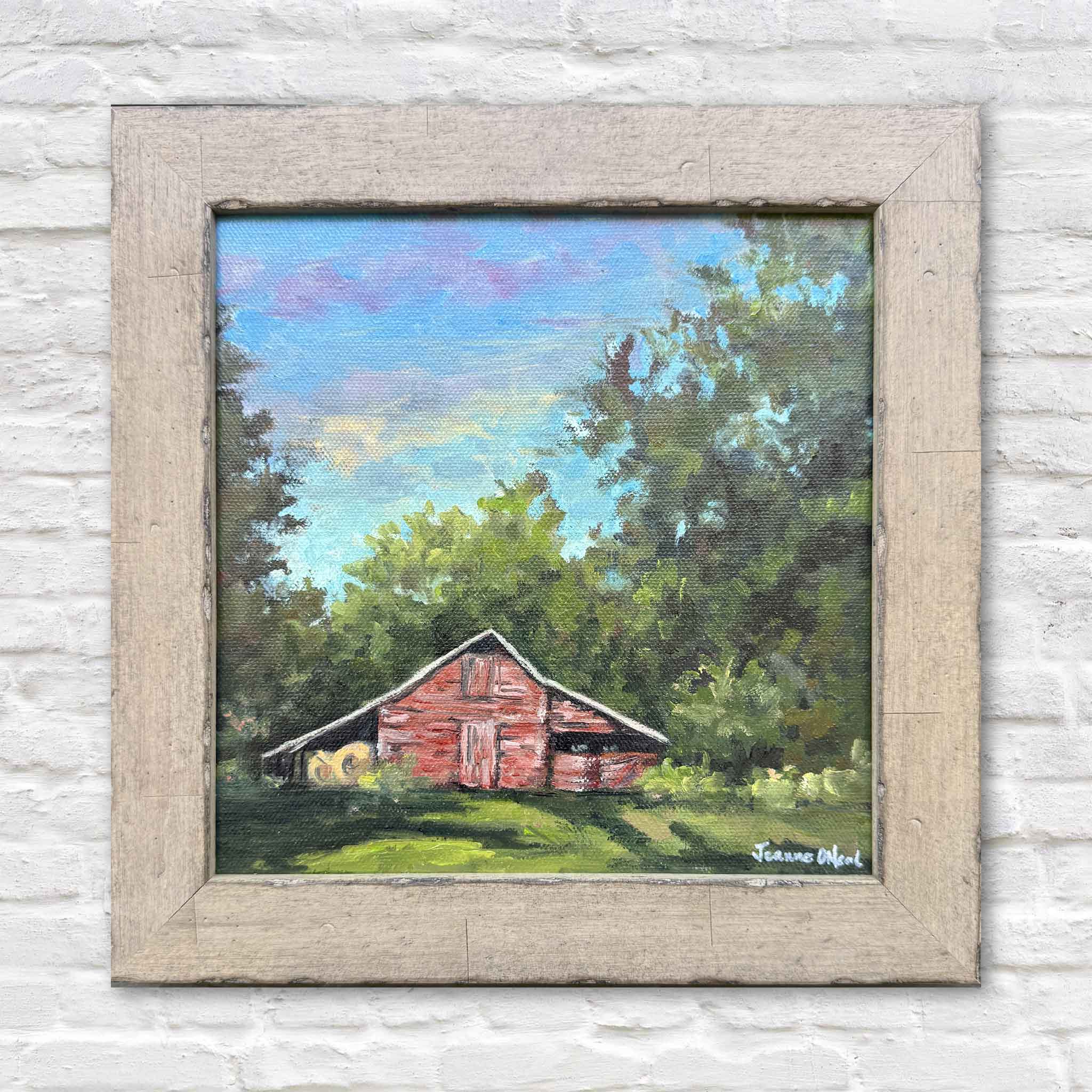 Original oil painting Grandaddy's Barn by Jeanne O'Neal featuring a weathered red barn surrounded by lush green trees beneath a soft blue and lavender sky. Framed artwork photographed on a white brick wall at Gray Man Gallery in Pawleys Island, South Carolina.