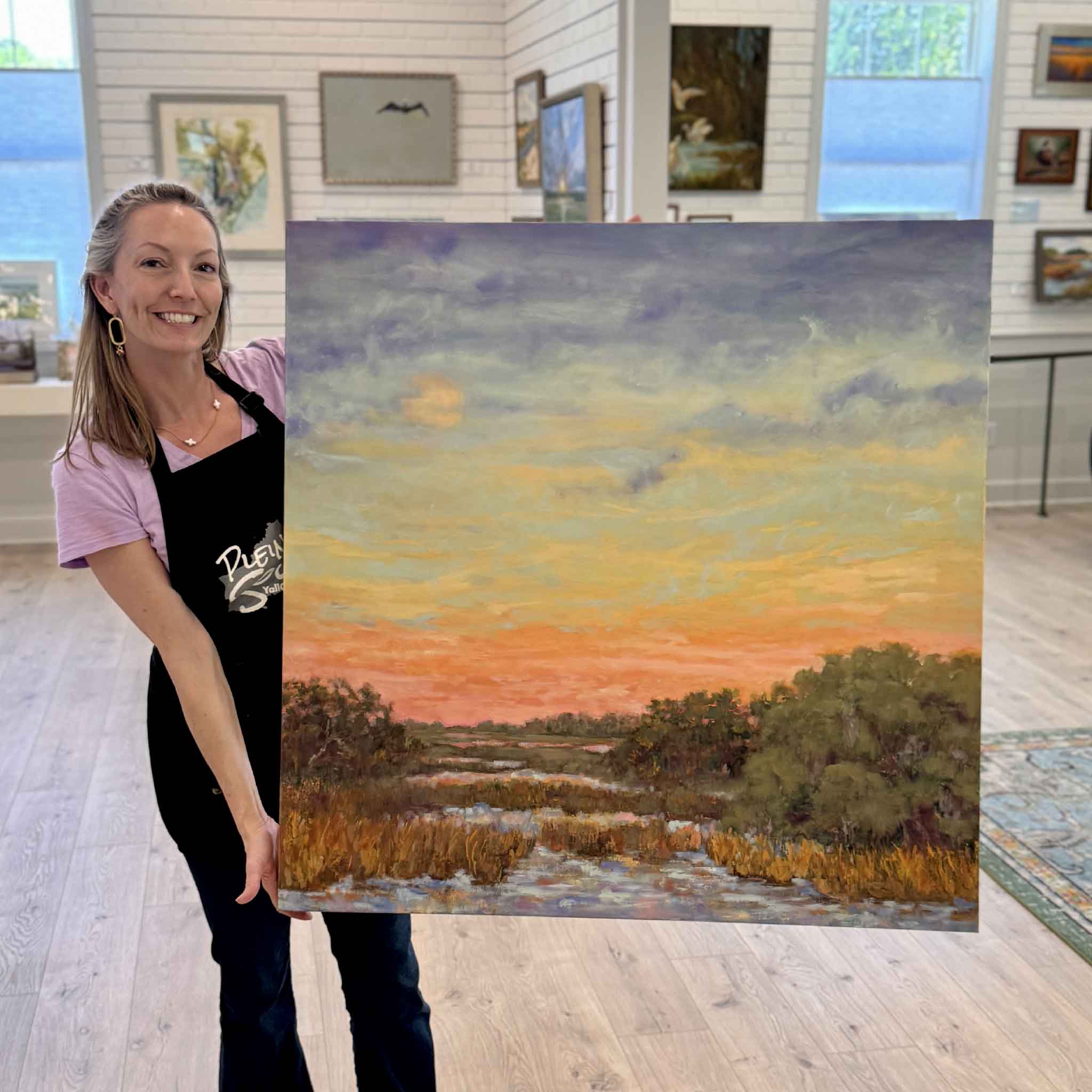 Jeanne O’Neal holding Hugs & Kisses original oil painting inside Gray Man Gallery Pawleys Island showing scale of gallery wrapped coastal marsh landscape at sunset