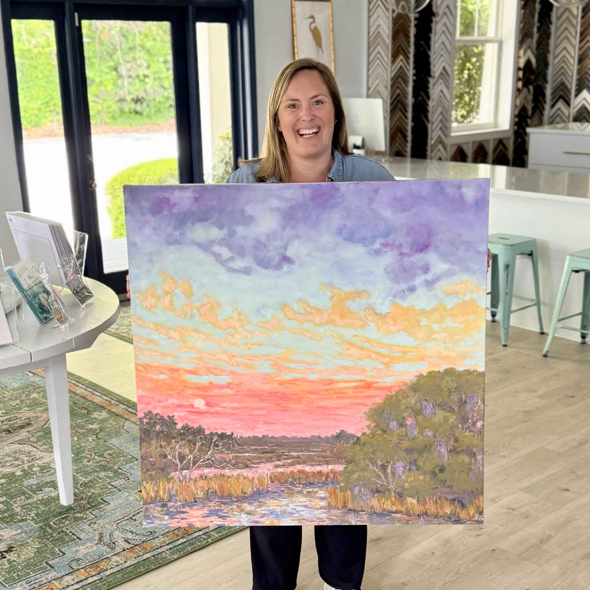 Bordy holding Hugs & Kisses Afterglow by Jeanne O’Neal inside Gray Man Gallery, a large original Lowcountry oil painting featuring a glowing sunset marsh with soft coral, peach, lavender, and blue skies reflected in winding water, showing scale of the artwork in a bright Pawleys Island gallery setting filled with local art.