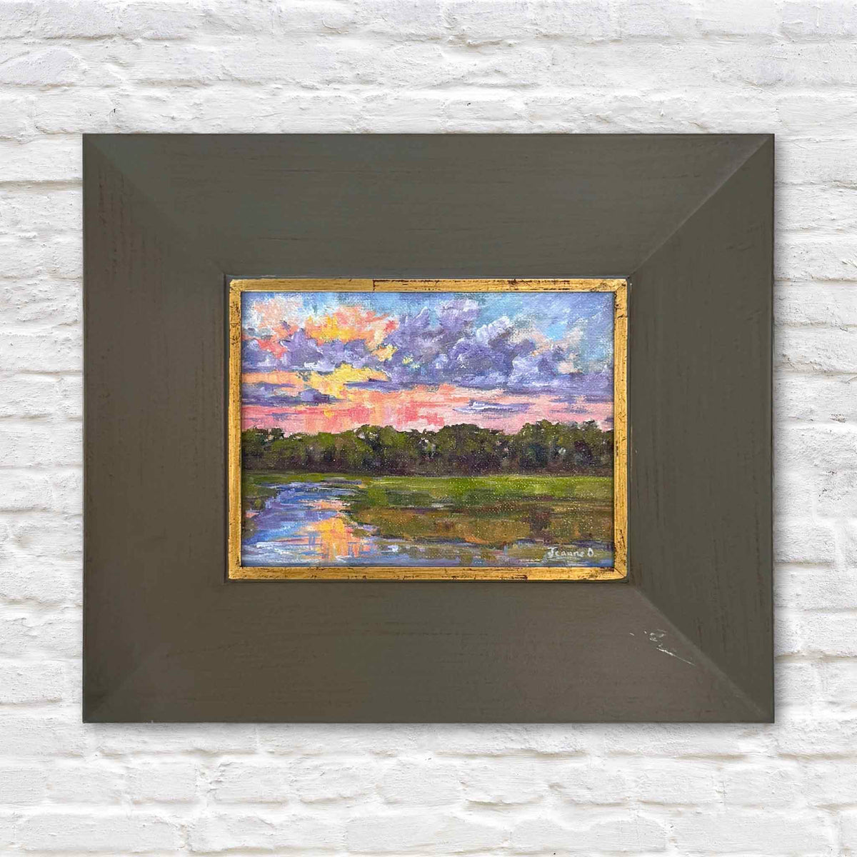 Lavender Haze, an original Lowcountry marsh sunset painting by local South Carolina artist Jeanne O'Neal, featuring dramatic lavender and violet clouds illuminated by brilliant coral, pink, and golden sunset light. A winding tidal creek reflects the colorful evening sky as it flows through lush green salt marsh grasses toward a distant tree-lined horizon. The small original coastal landscape is presented in a wide wood frame with a distressed gold inner border, at Gray Man Gallery in Pawleys Island.
