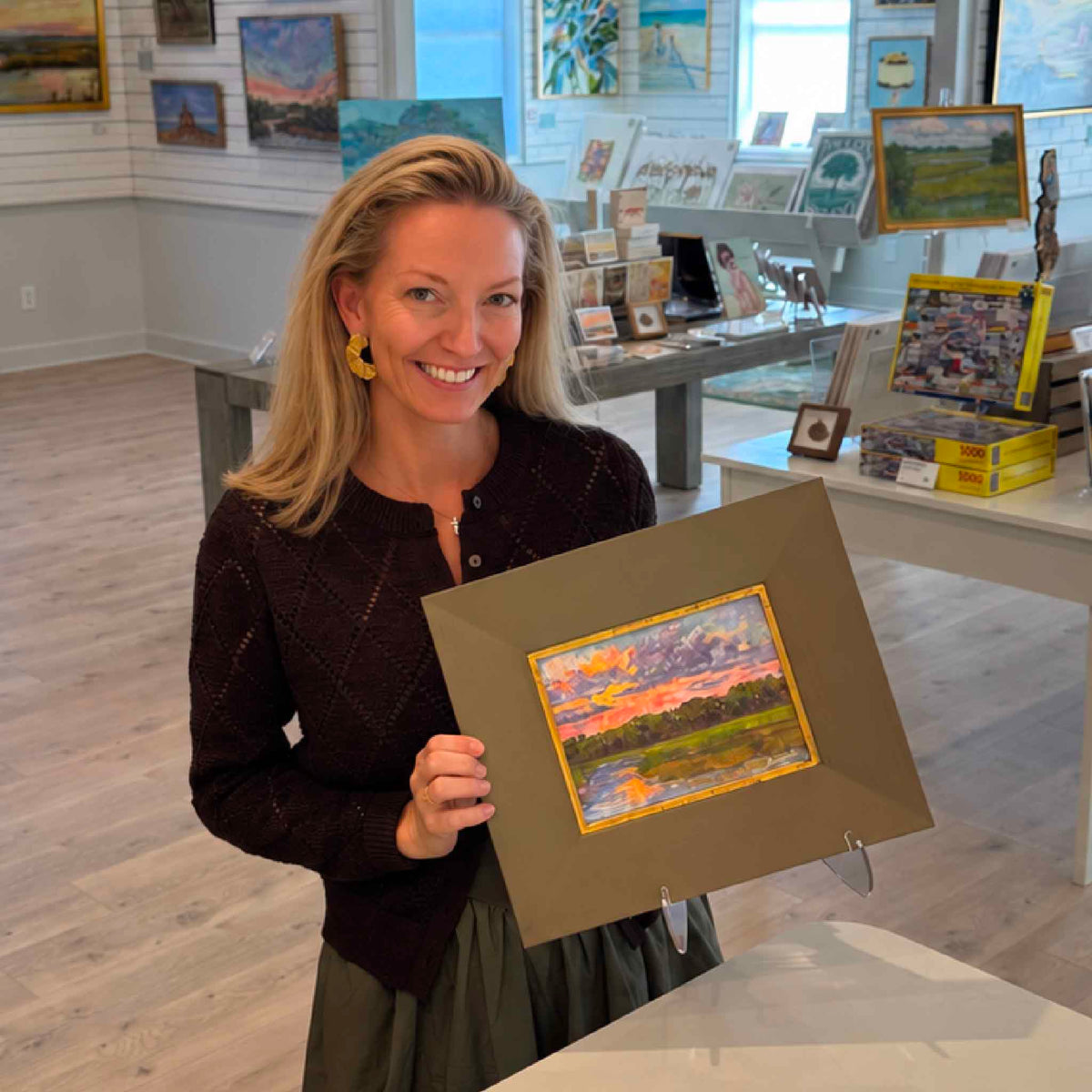 Gray Man Gallery team member holding Lavender Haze, a small original framed Lowcountry landscape painting by South Carolina artist Jeanne O'Neal, inside Gray Man Gallery in Pawleys Island. The colorful artwork depicts a winding tidal creek, green coastal marsh grasses, and a dramatic sunset sky filled with lavender, purple, coral, and golden clouds. The painting is surrounded by a substantial charcoal-gray wood frame with a distressed gold inner border, demonstrating its scale and presentation.