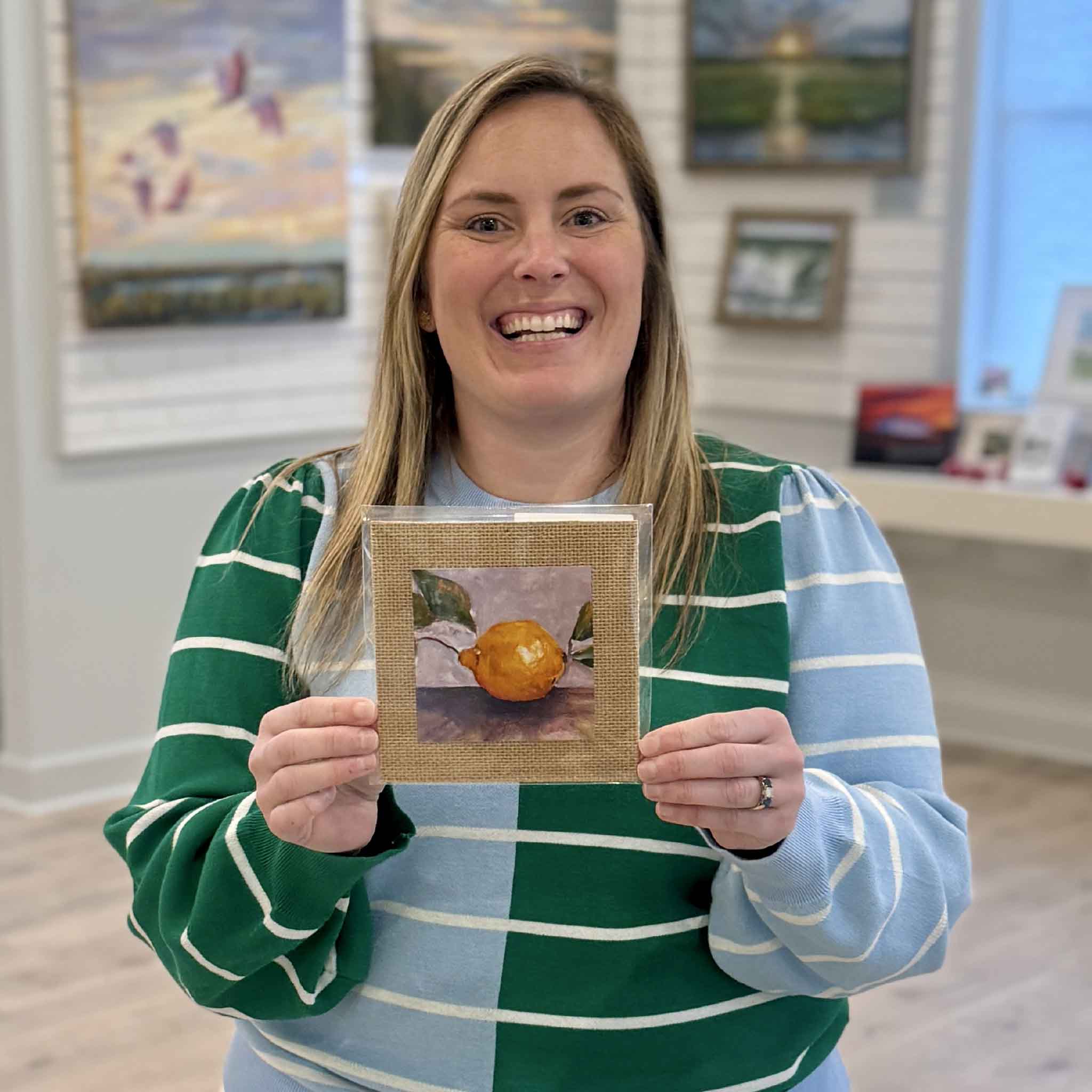 A team member at Gray Man Gallery in Pawleys Island, South Carolina holds Jeanne O’Neal’s “Main Squeeze” lemon print, a 4x4 inch artwork mounted on a 6x6 textured burlap mat. The image highlights the scale, vibrant citrus color palette, and coastal charm of this Lowcountry local artist print, available exclusively at Gray Man Gallery.