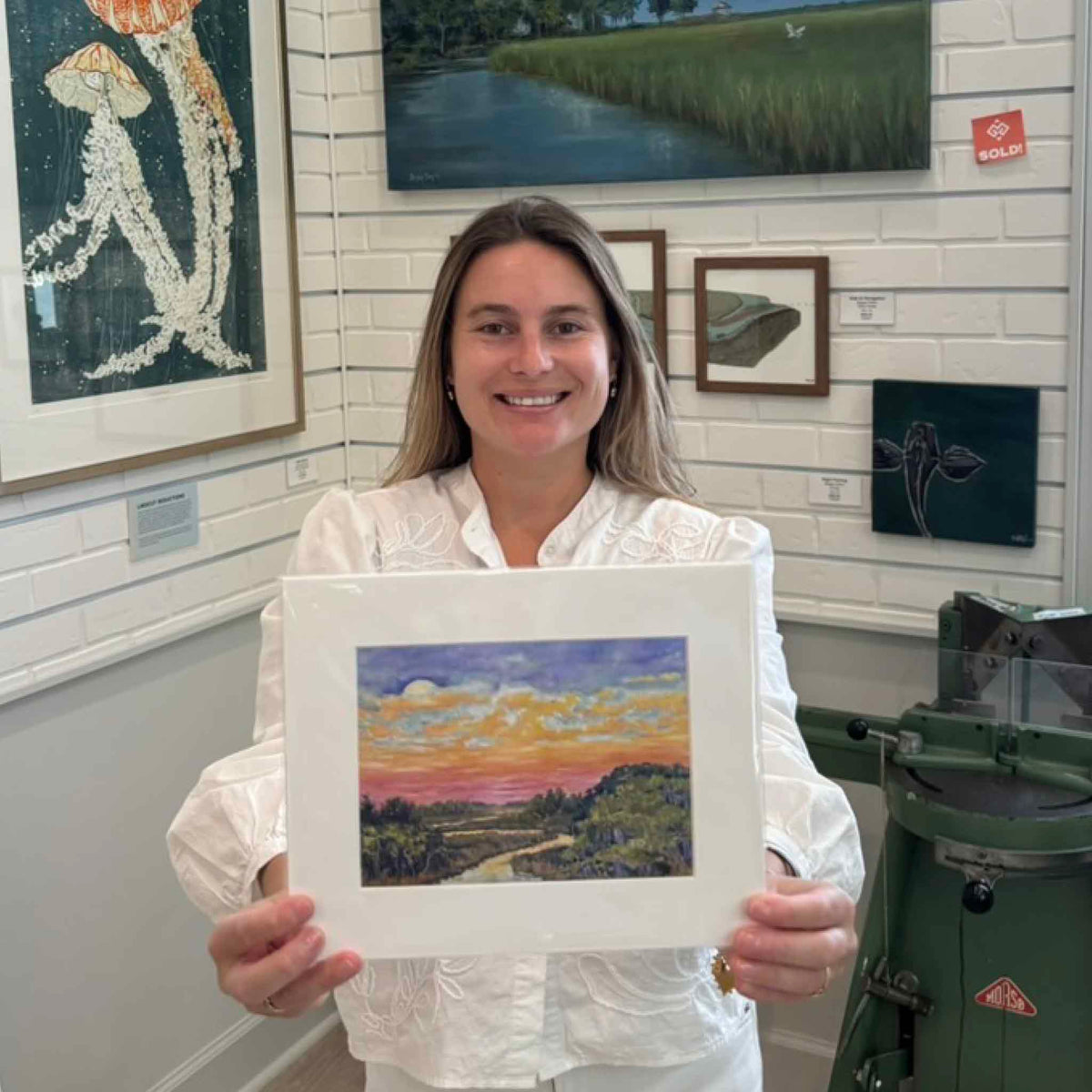 Kyra holding the larger standard-size Moonrise fine art giclée print by Jeanne O’Neal inside Gray Man Gallery in Pawleys Island, South Carolina, showing the vibrant coastal marsh landscape and moonlit evening sky.