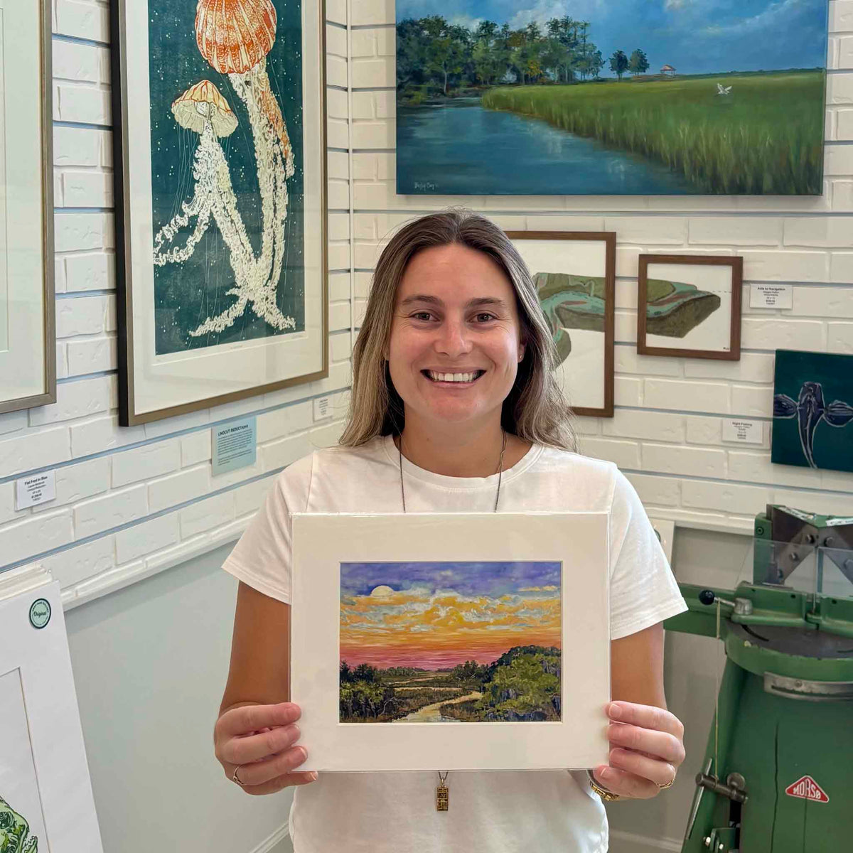 Kyra holding the smaller standard-size Moonrise fine art giclée print by Jeanne O’Neal inside Gray Man Gallery in Pawleys Island, South Carolina, featuring a moon rising over a colorful Lowcountry marsh and winding tidal creek.
