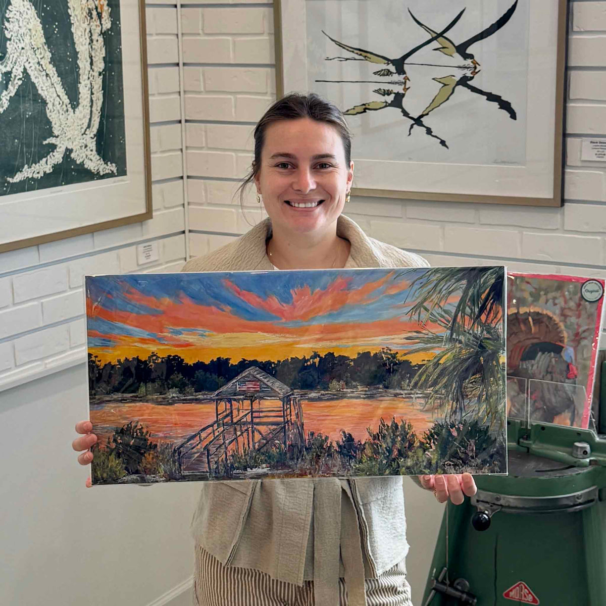 Gray Man Gallery team member holding Shabby Dock, a colorful panoramic fine art giclée print by local South Carolina artist Jeanne O'Neal, inside Gray Man Gallery in Pawleys Island. The wide-format coastal landscape features a rustic covered dock with an American flag, glowing orange sunset reflections, vibrant pink and blue clouds, dark shoreline trees, and palmetto fronds, with the photograph demonstrating the print's panoramic proportions and display scale.