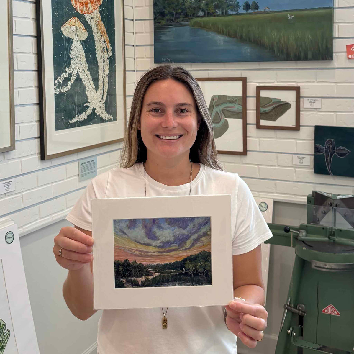 Kyra holding Jeanne O’Neal’s The Day Softens fine art giclée print inside Gray Man Gallery in Pawleys Island, South Carolina, featuring a colorful coastal marsh and winding tidal creek at sunset, professionally matted to fit a standard-size frame.