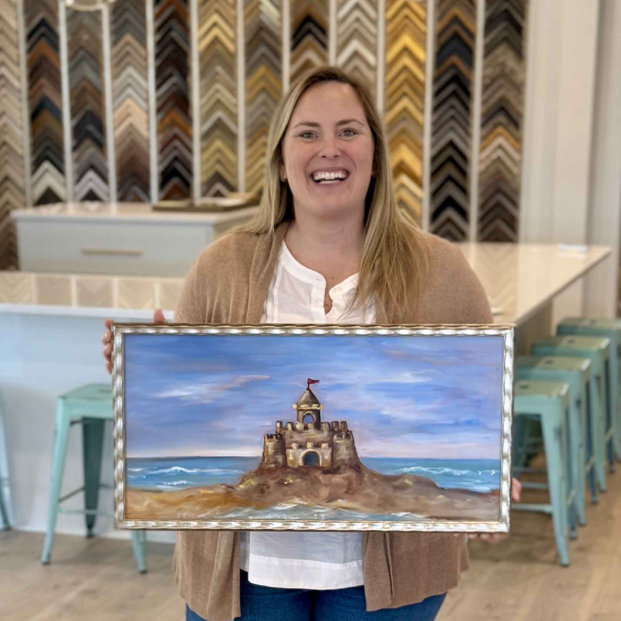 Local Pawleys Island artist Jenny Reynolds holding her original oil painting Castle in the Sand inside Gray Man Gallery, featuring a whimsical coastal sandcastle scene in her signature champagne frame
