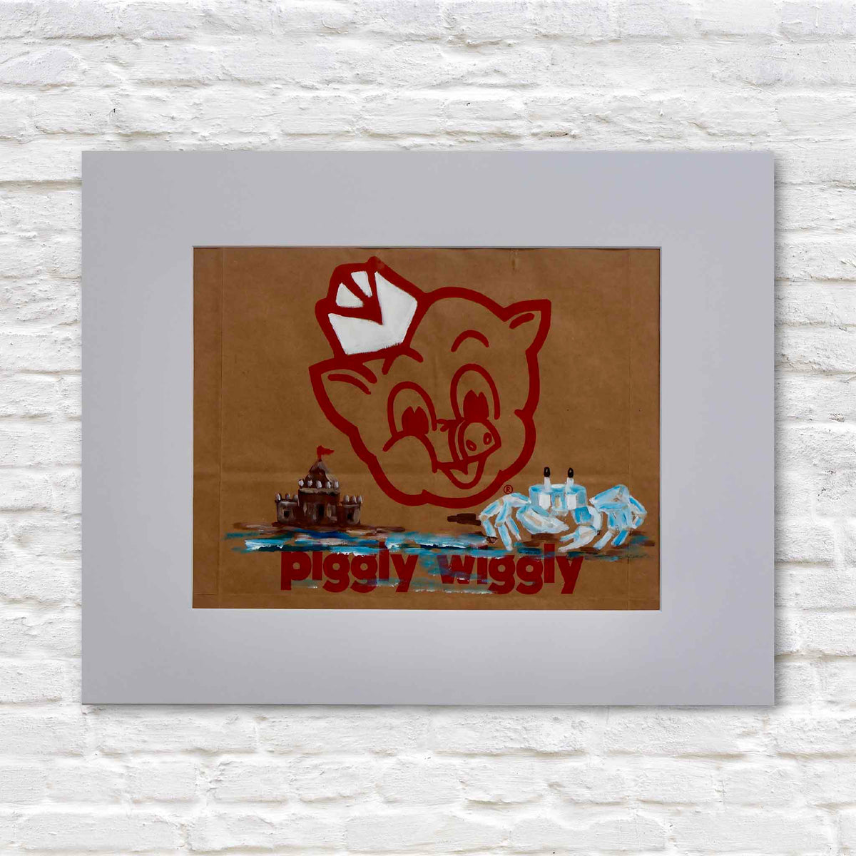 King of the Castle original Piggly Wiggly grocery bag artwork by local artist Jenny Reynolds, featuring a hand-painted blue ghost crab, sandcastle and coastal water beneath the vintage red pig logo. Presented in a white mat against a white brick background at Gray Man Gallery in Pawleys Island, South Carolina.