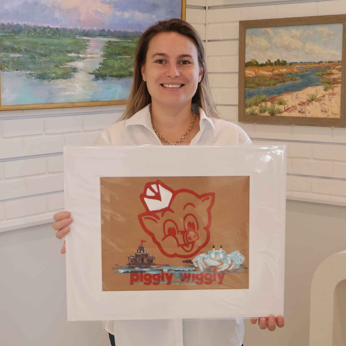 Gray Man Gallery team member holding King of the Castle by local artist Jenny Reynolds, an original coastal painting on a vintage Piggly Wiggly paper grocery bag featuring a blue ghost crab and sandcastle. The artwork is displayed with a wide white mat inside Gray Man Gallery in Pawleys Island, South Carolina.