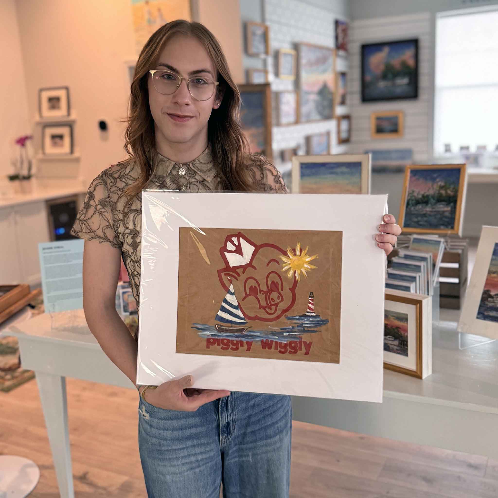 Alex holding “Piggly Wiggly: Sail Away,” an original Jenny Reynolds painting on a Piggly Wiggly grocery bag featuring sailboats, water, and a bright sun.