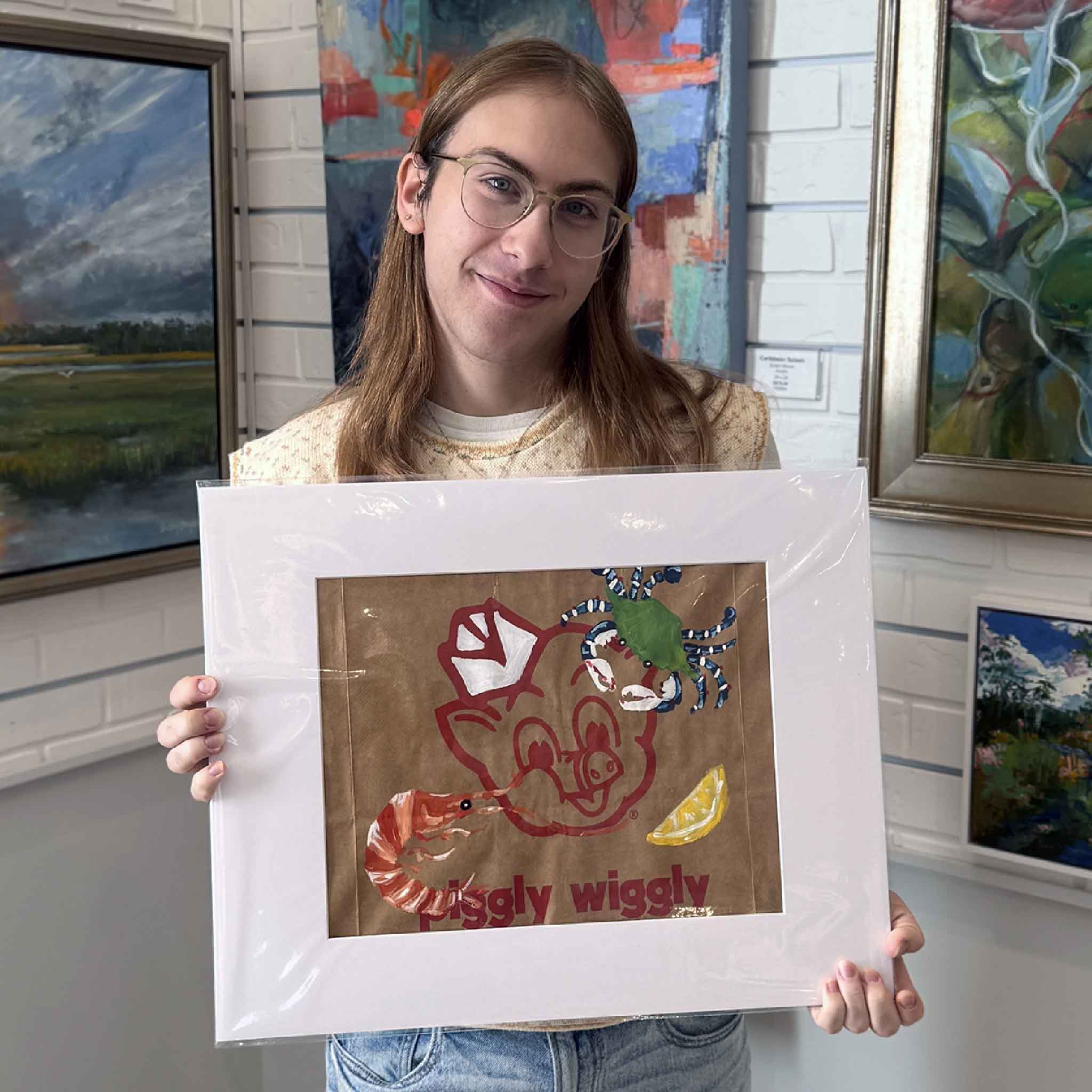 Alex holding local artist Jenny Reynolds's original hand-painted Piggly Wiggly grocery bag artwork titled Shrimp, Crab & Lemon at Gray Man Gallery in Pawleys Island, SC. This one-of-a-kind Lowcountry piece features the classic pig mascot with coastal seafood imagery and is matted to a standard 16x20 size, making it an easy-to-frame Southern art gift.