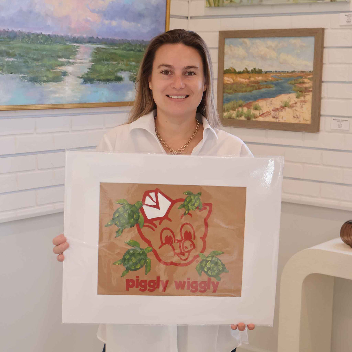 Gray Man Gallery team member holding The Great Escape by local artist Jenny Reynolds, an original acrylic painting of four green sea turtles on an authentic Piggly Wiggly grocery bag. The artwork is displayed in a wide white mat inside the Pawleys Island, South Carolina art gallery.