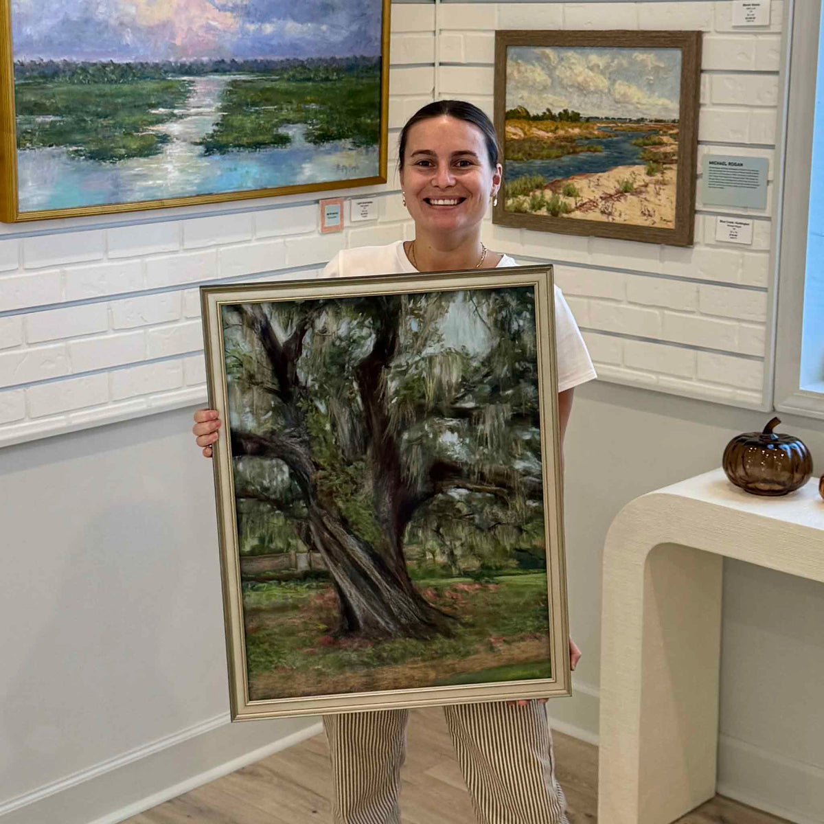 Gray Man Gallery team member holding Nature's Majesty, an original framed pastel painting by local artist Joanne Willoughby, inside the gallery in Pawleys Island, South Carolina. The vertical landscape depicts a majestic live oak at Brookgreen Gardens, with deeply textured bark, wide twisting limbs, hanging Spanish moss, and a lush green setting, showing the scale and elegant champagne-gold framing of the original artwork.