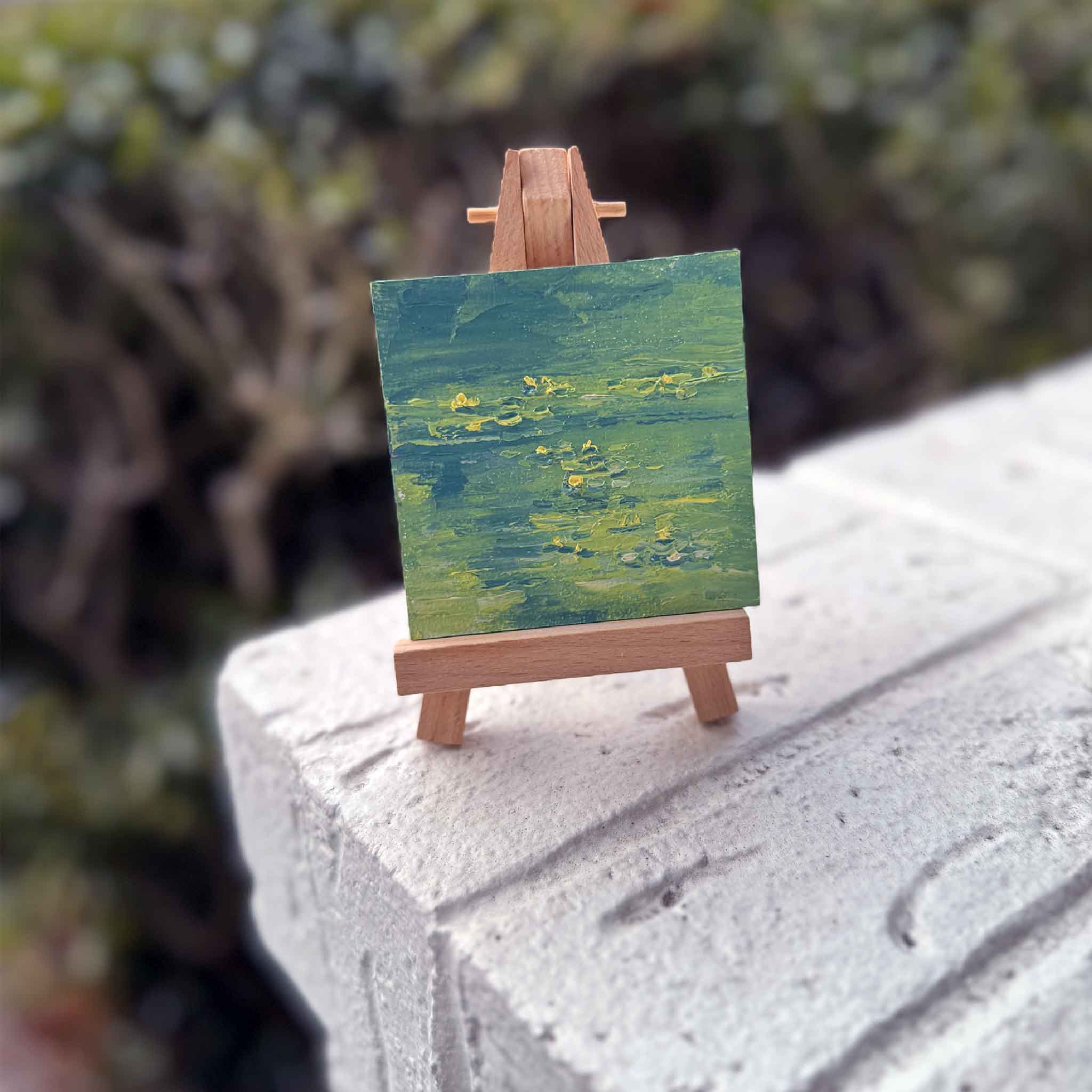 Original miniature oil painting of a lily pond with soft green and blue water and small yellow blooms, displayed on a tiny wooden easel outdoors at Gray Man Gallery.