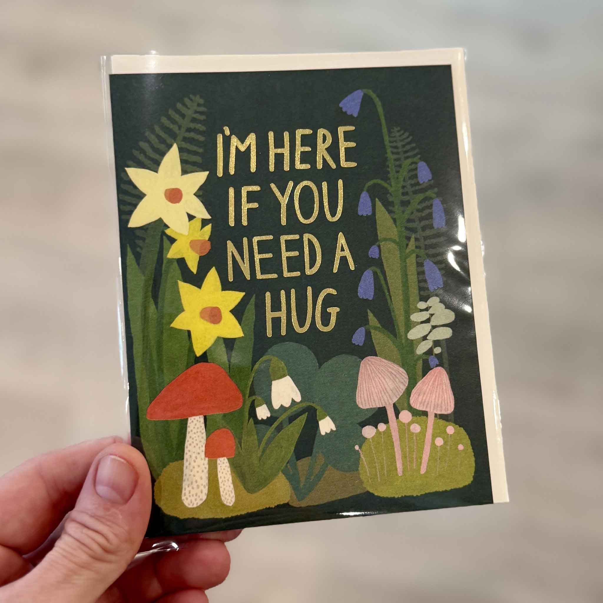 Garden Hugs encouragement greeting card illustrated by Kate Pugsley for Red Cap Cards, photographed inside Gray Man Gallery in Pawleys Island, South Carolina. This premium blank-inside greeting card features gold foil lettering surrounded by illustrated daffodils, bluebells, mushrooms, and woodland plants with the heartfelt message "I'm Here If You Need a Hug."