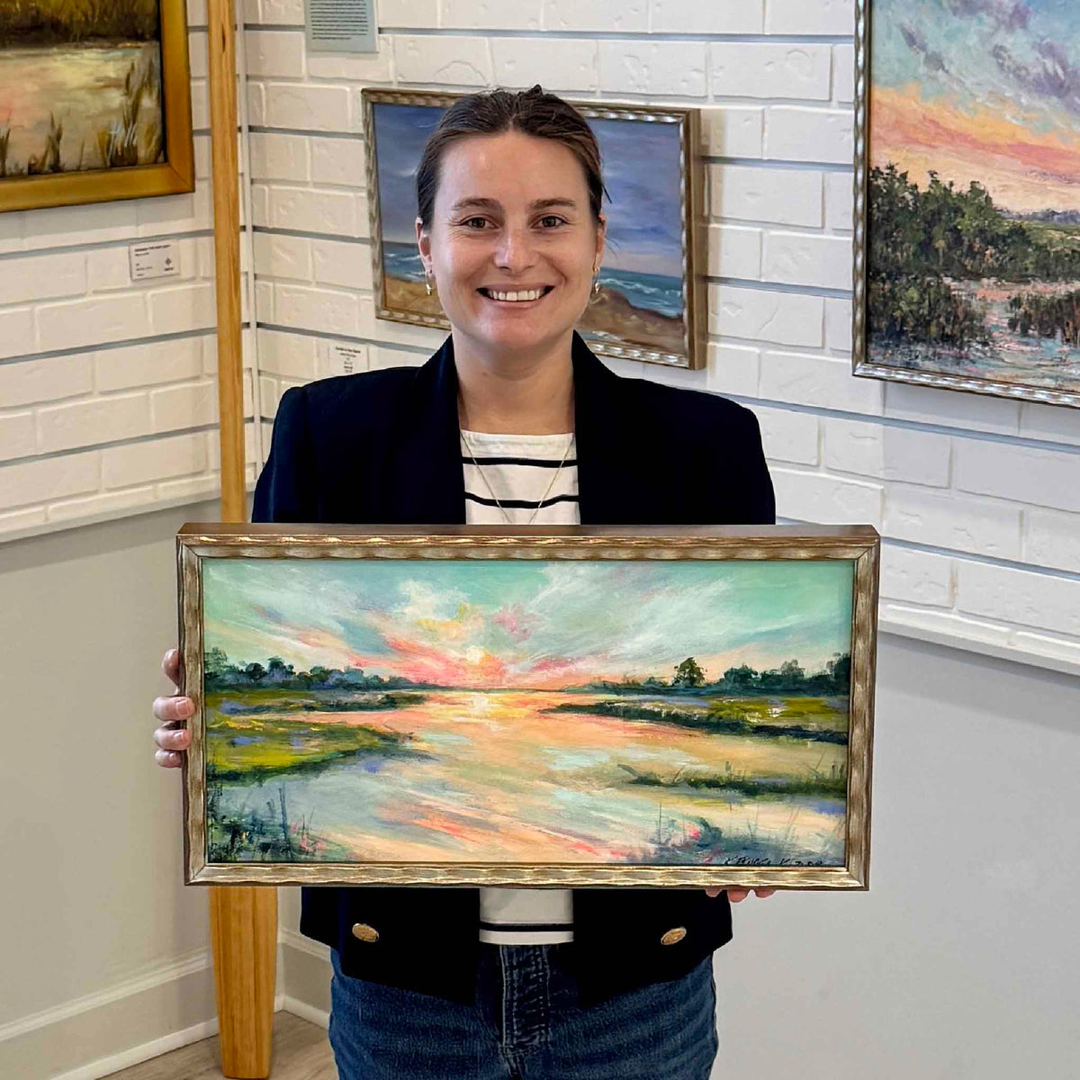 Gray Man Gallery team member holding The Last Blush, an original oil painting by local artist Kathleen Kazior, inside Gray Man Gallery in Pawleys Island, South Carolina. The horizontal Lowcountry landscape features a vivid coral and turquoise sunset reflecting across a winding tidal creek, framed in distressed metallic gold.