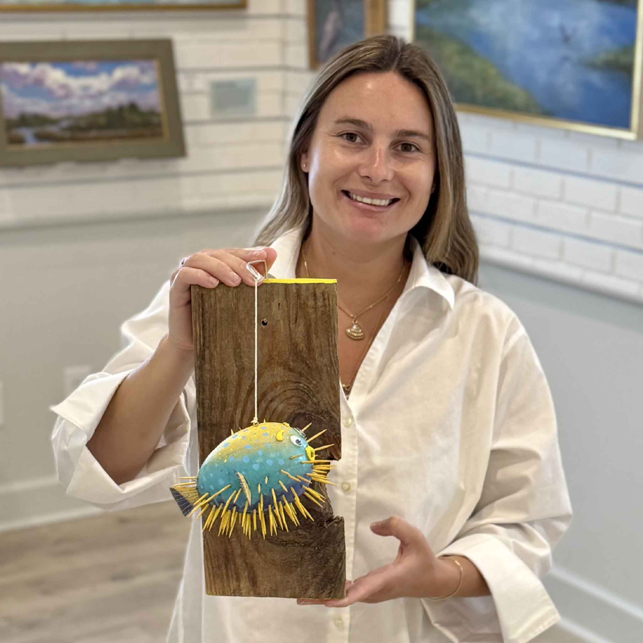Kyra holding Pufferfish #18, a one-of-a-kind mixed media wall sculpture by Charleston artist Nick Kawczynski made from papier-mâché, reclaimed wood, toothpicks, acrylic paint, and found materials at Gray Man Gallery.