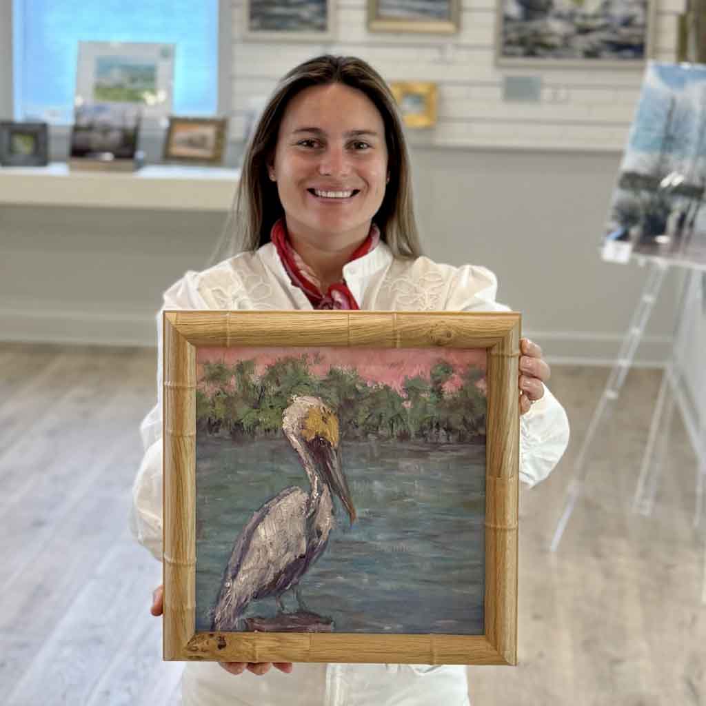 Kyra holding A Moment Alone, an original pelican oil painting by Jeanne O'Neal featuring soft coastal colors, tranquil marsh waters, and a handcrafted bamboo frame at Gray Man Gallery.