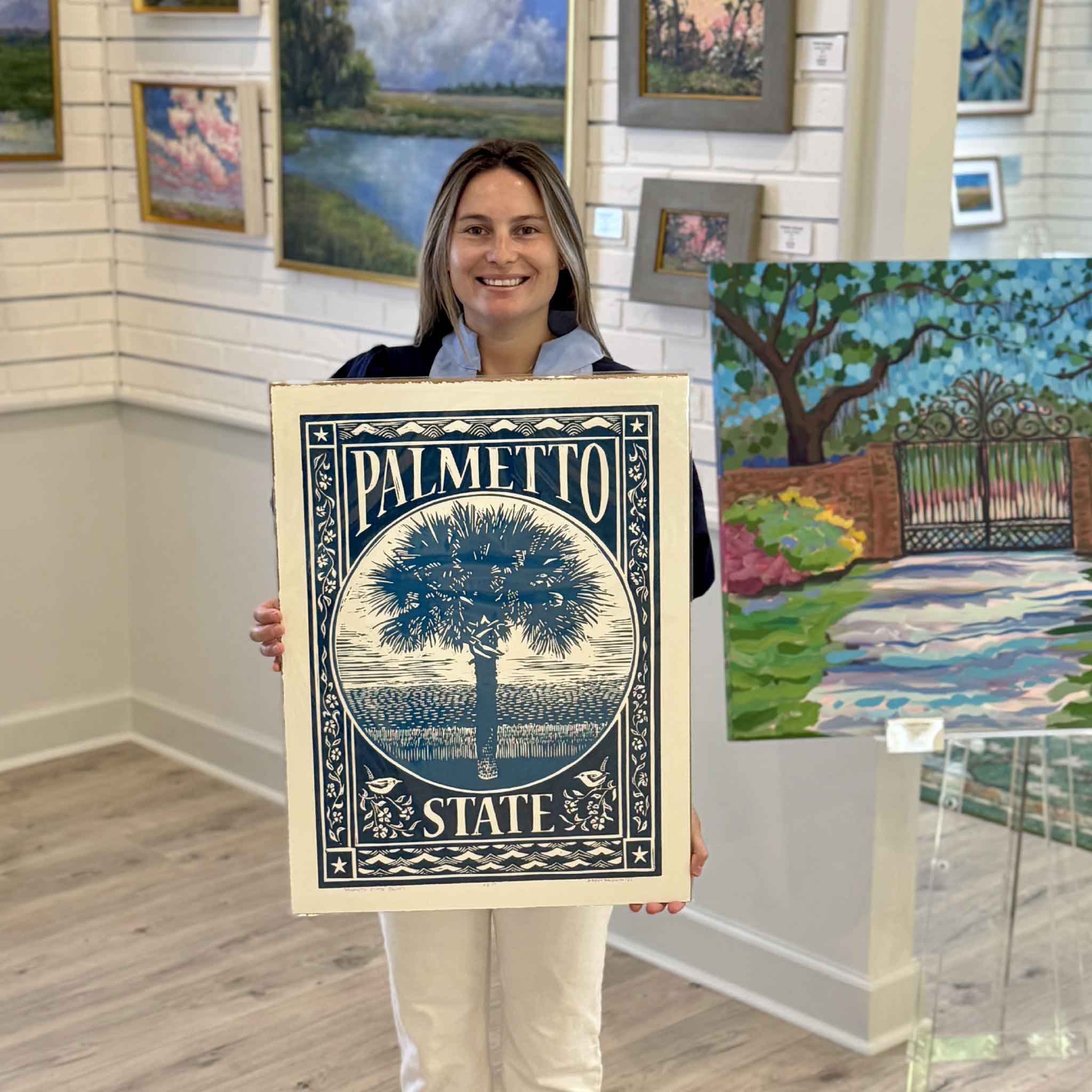 Kyra holding Palmetto State, an original hand-pulled linocut by Aaron Baldwin featuring a blue palmetto tree, coastal marsh landscape, and decorative South Carolina motifs. Photographed inside Gray Man Gallery in Pawleys Island, South Carolina.